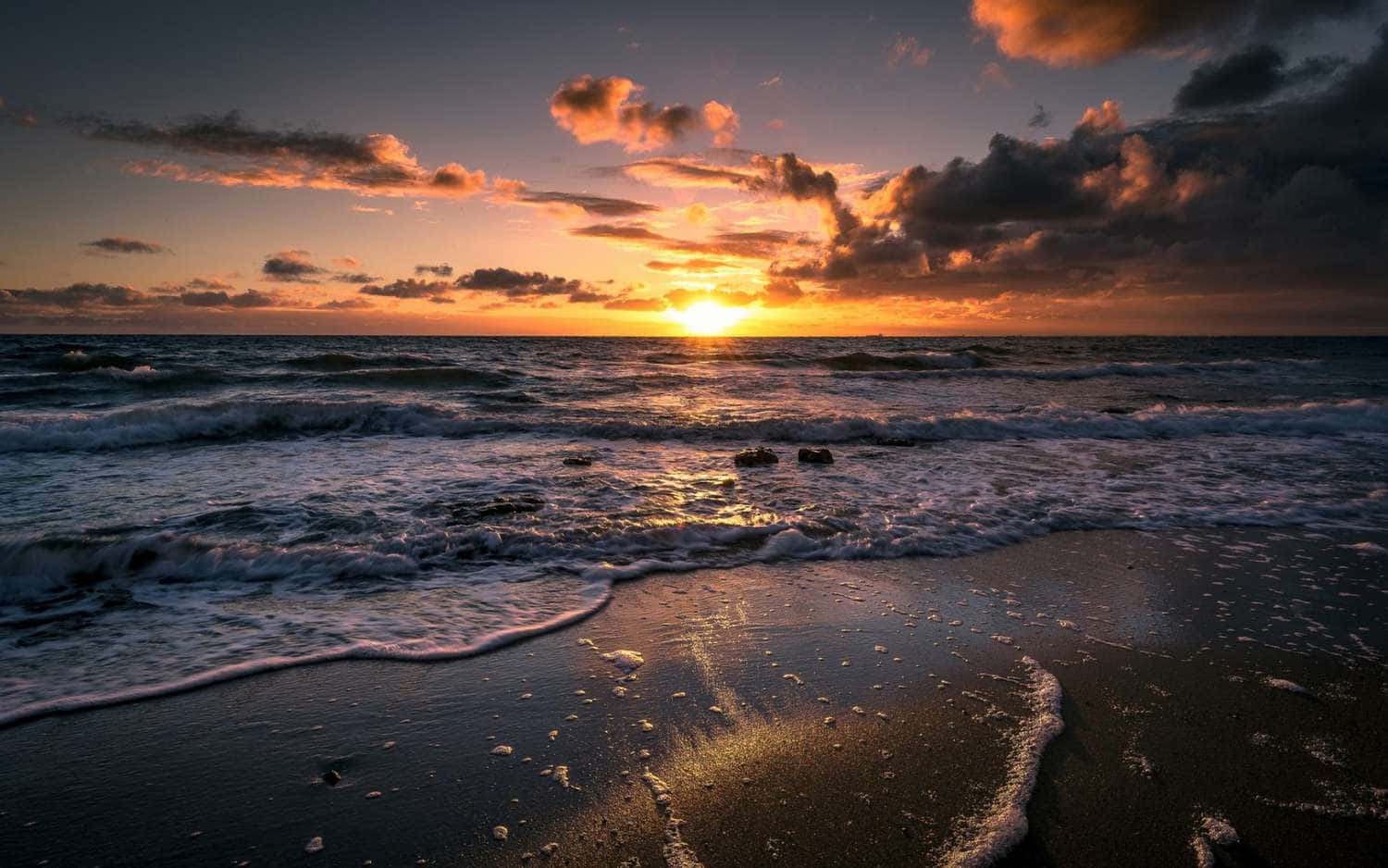 Sea 桌布 壁紙 — 夕陽將天空染成火熱的橙色和紫色，海浪拍打著深色沙灘。