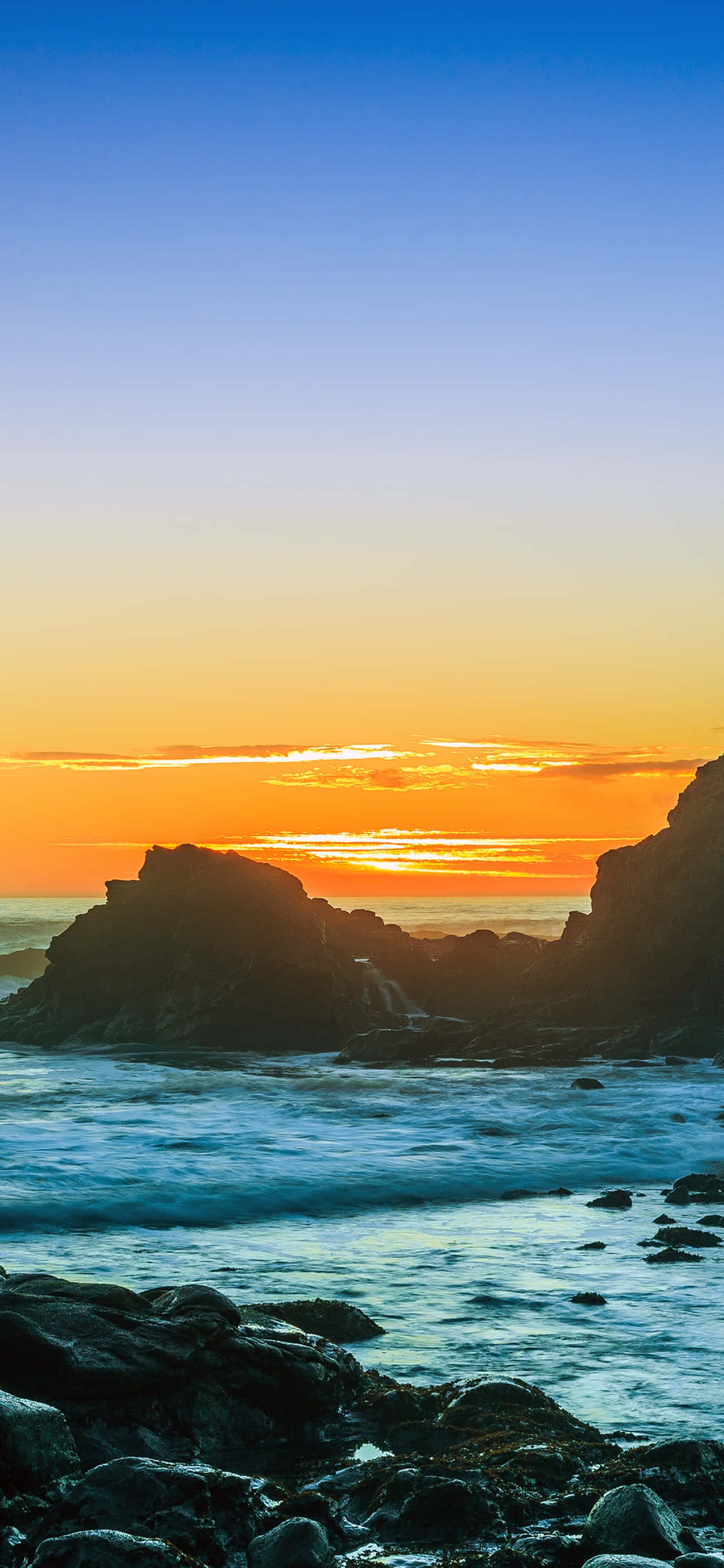 iPhone X Malibu 桌布 壁紙 — 夕陽將天空染成火熱的橙色和藍色，海浪拍打著深色的海岸岩石。