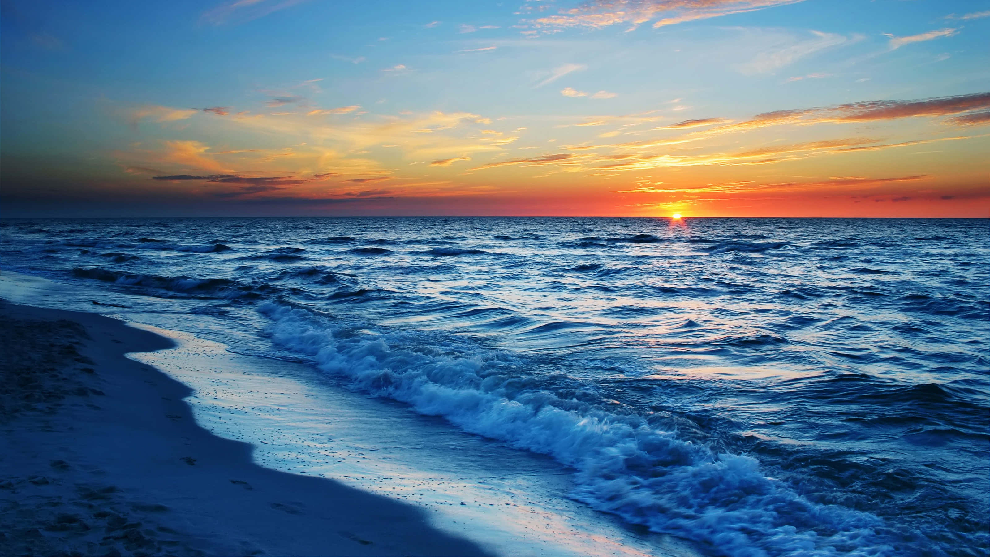 Calm 桌布 壁紙 — 夕陽將天空染成火熱的橙色和藍色，海浪拍打著沙灘。