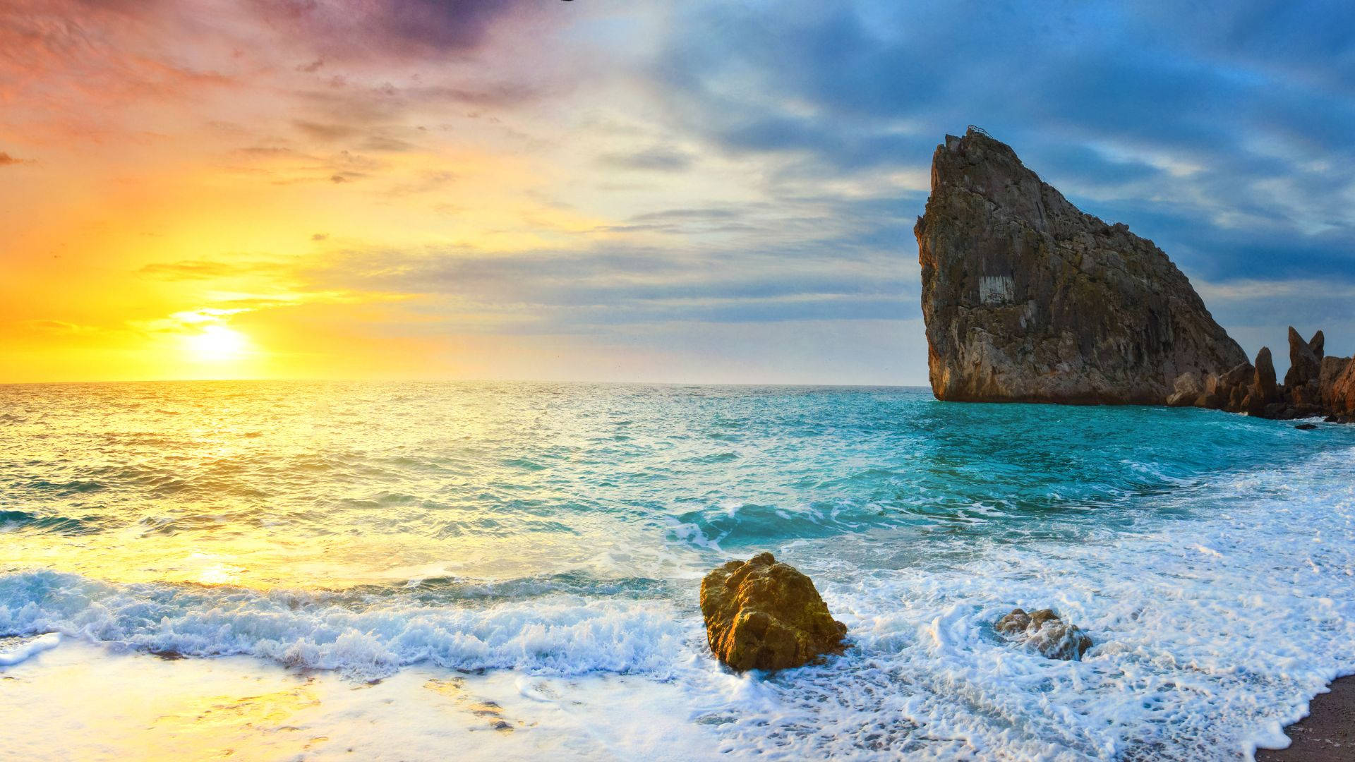 海灘 桌布 壁紙 — 夕陽將天空染成火熱的橙色和藍色，海浪拍打著寧靜海灘上的岩石。