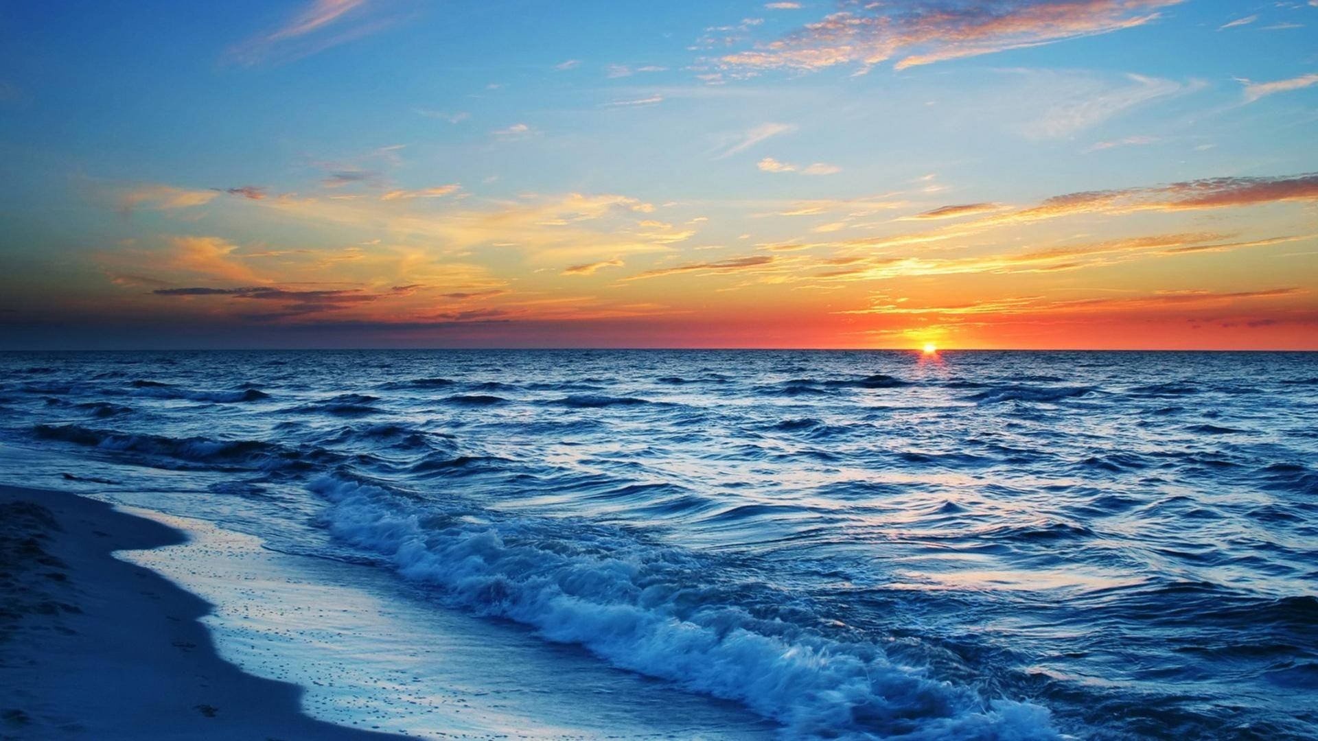 夕陽天空 桌布 壁紙 — 夕陽將天空染成火熱的橙色和藍色，海浪拍打著沙灘。