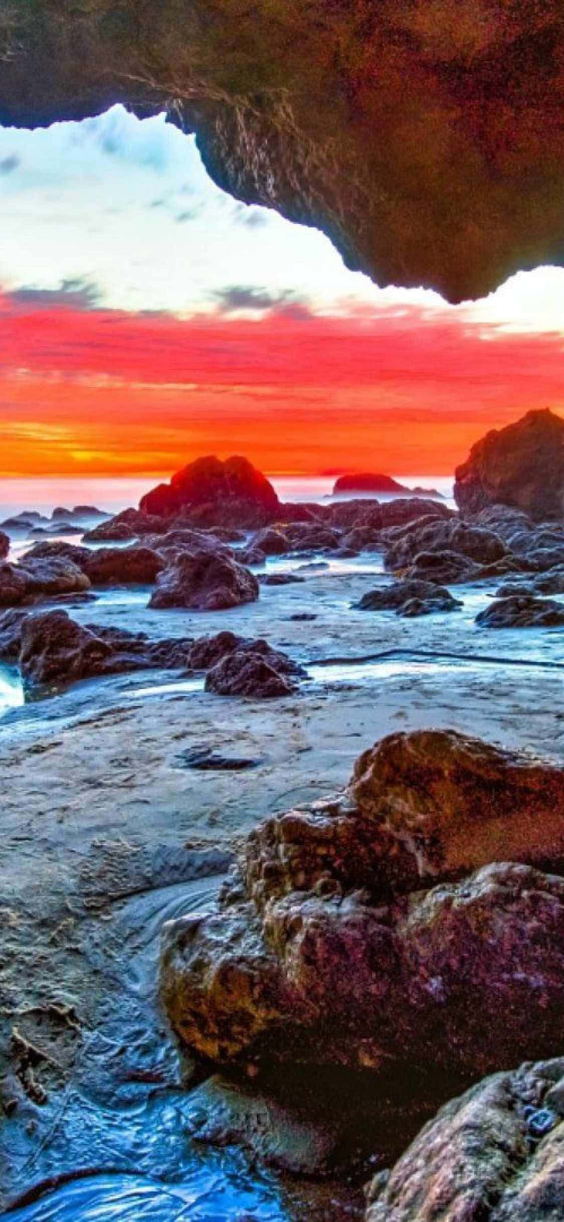iPhone Xs Malibu 桌布 壁紙 — 夕陽將天空染成火紅和橙色，海浪輕輕拍打天然岩石拱門下的崎嶇海岸岩石。