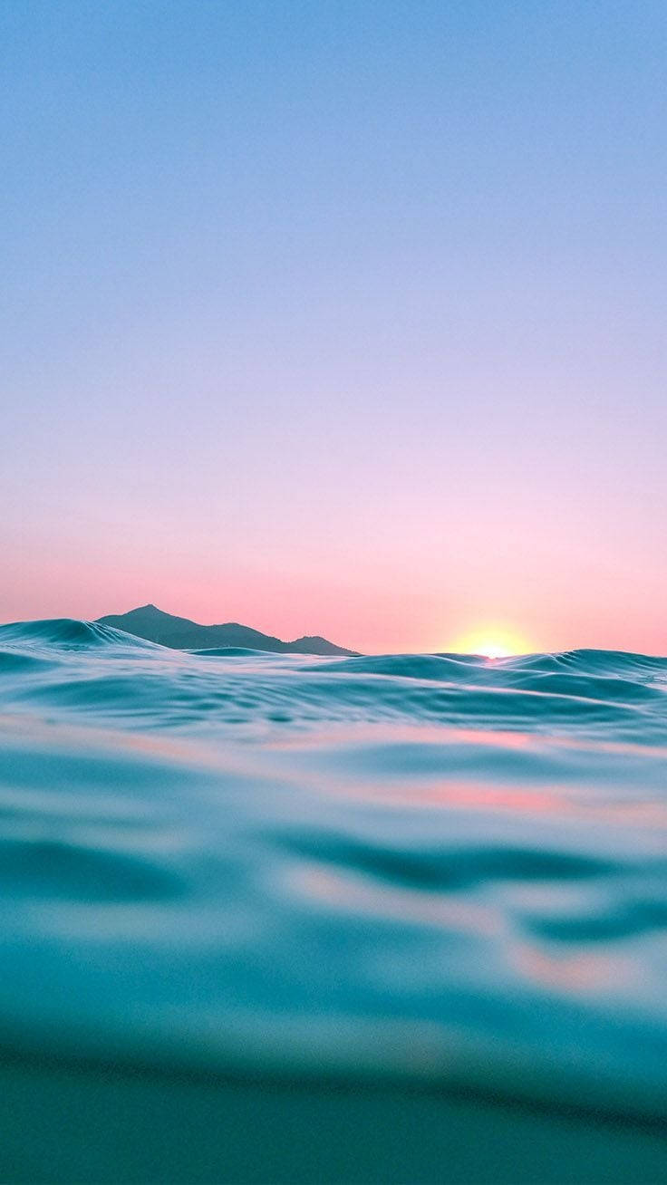 海洋 桌布 壁紙 — 夕陽將天空染成粉紅色和藍色，映照在遠山之上，溫柔的海浪在前景中泛起漣漪。
