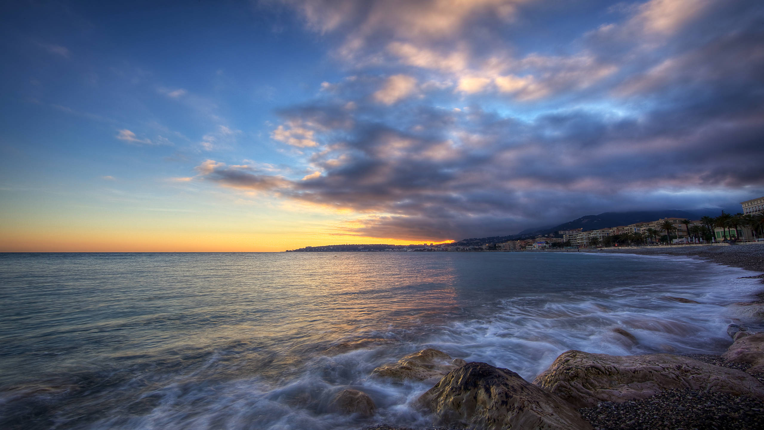 Monaco 桌布 壁紙 — 夕陽將天空染成金橙色和深藍色，海浪拍打著海邊小鎮旁的岩石海岸。