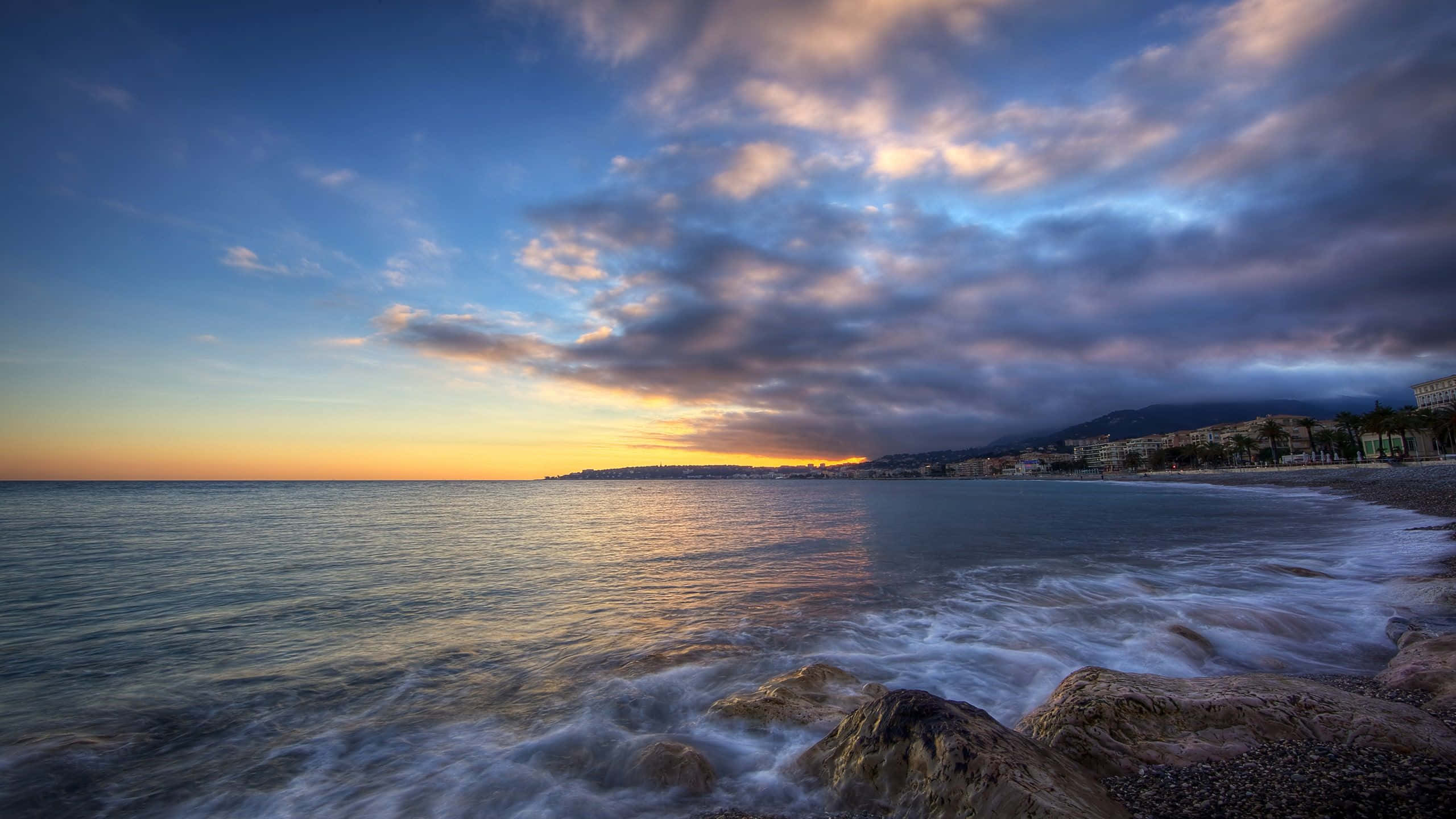 Coastline 桌布 壁紙 — 夕陽將天空染成金橙色和深藍色，海浪拍打岩石海岸，遠處有沿海小鎮。