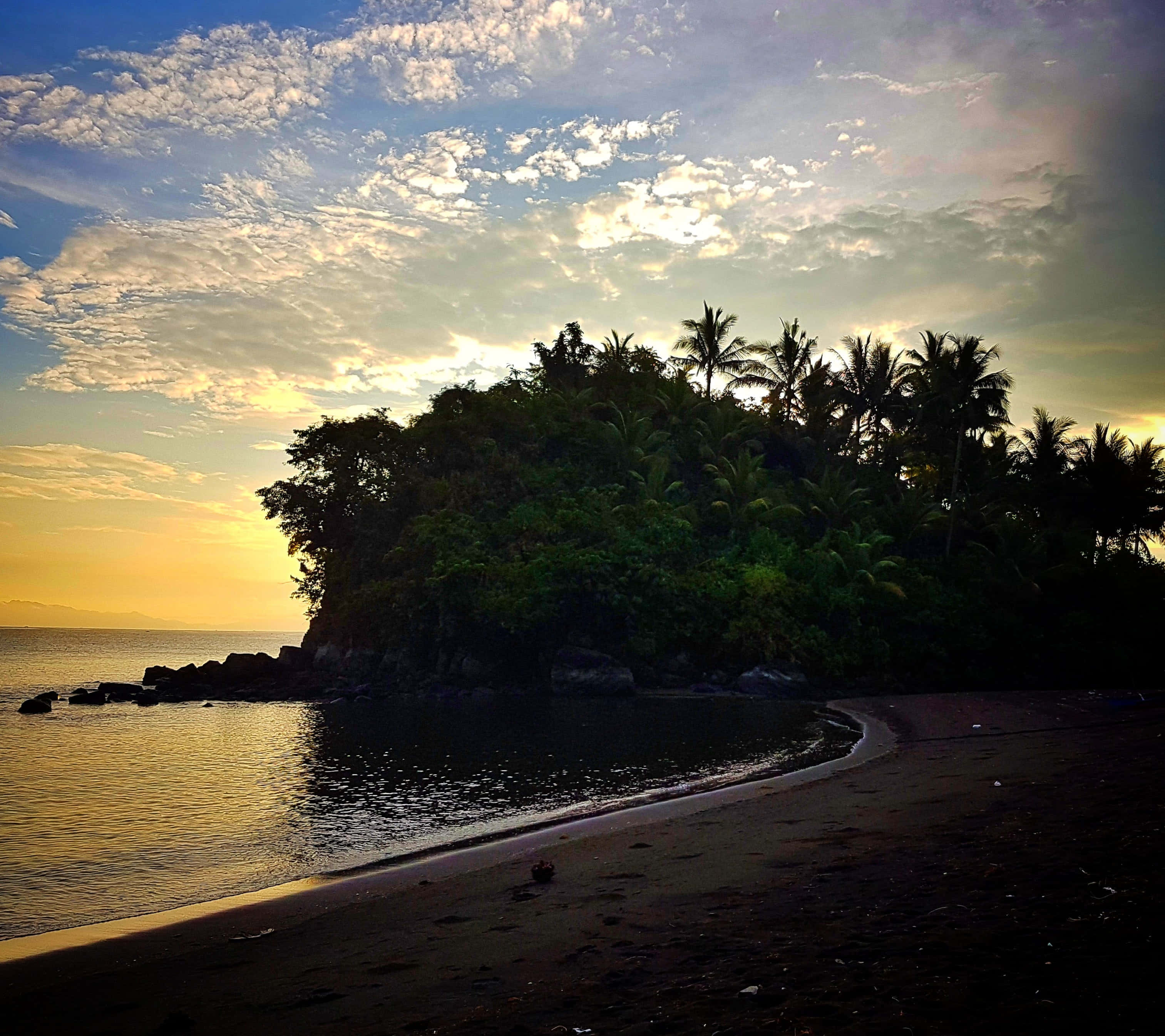 Dark Island 桌布 壁紙 — 夕陽將天空染成金色，海浪拍打著棕櫚樹環繞的茂密島嶼旁的深色沙灘。