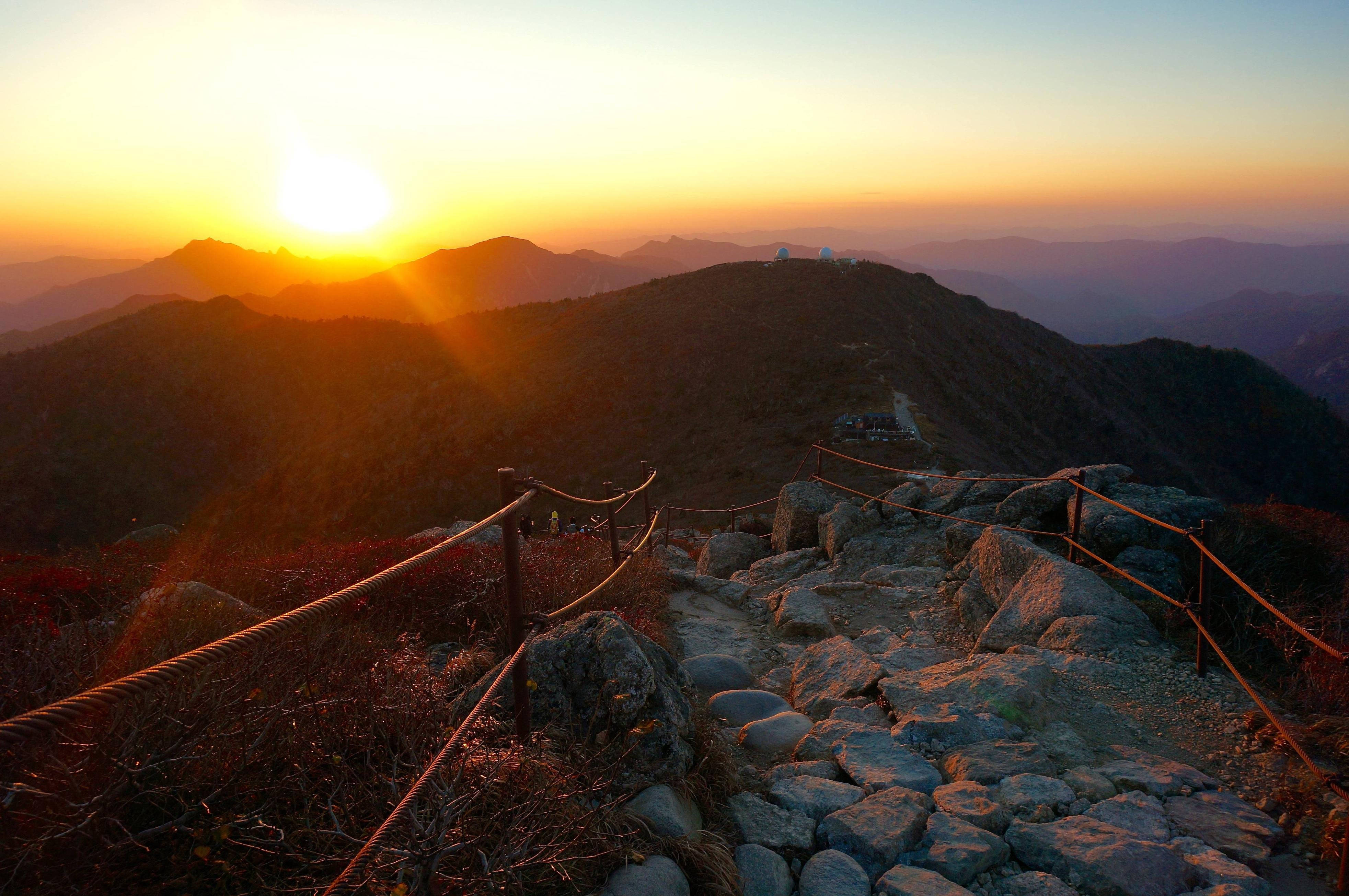 山脈 桌布 壁紙 — 夕陽將崎嶇的山間小徑沐浴在溫暖的金色光芒中，遠方的山峰逐漸消失在朦朧的暮色中。