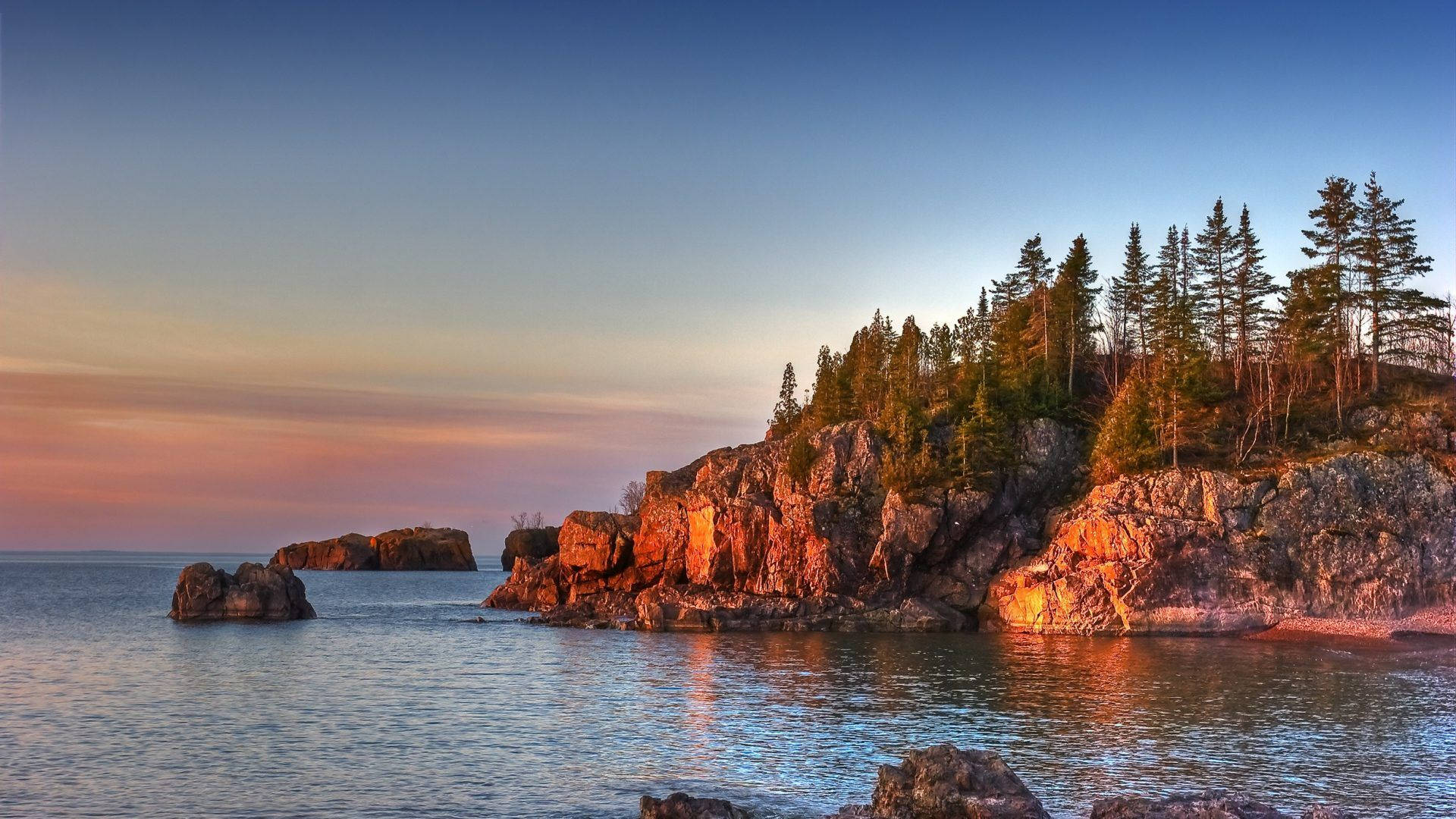 Minnesota 桌布 壁紙 — 夕陽將崎嶇的海岸懸崖和松樹沐浴在溫暖的橙色光芒中，倒映在平靜的水面上。