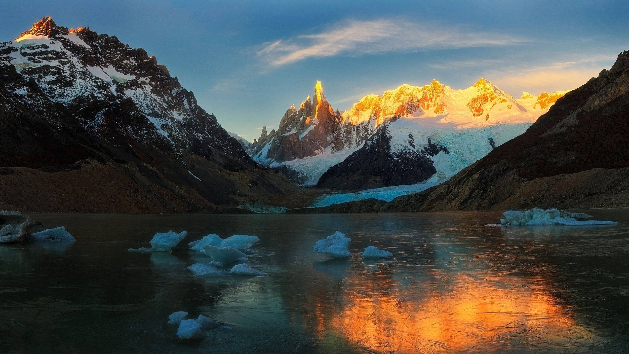 Argentina 桌布 壁紙 — 夕陽將崎嶇的雪山沐浴在金色光芒中，反射在結冰的湖面上，湖中漂浮著冰山。