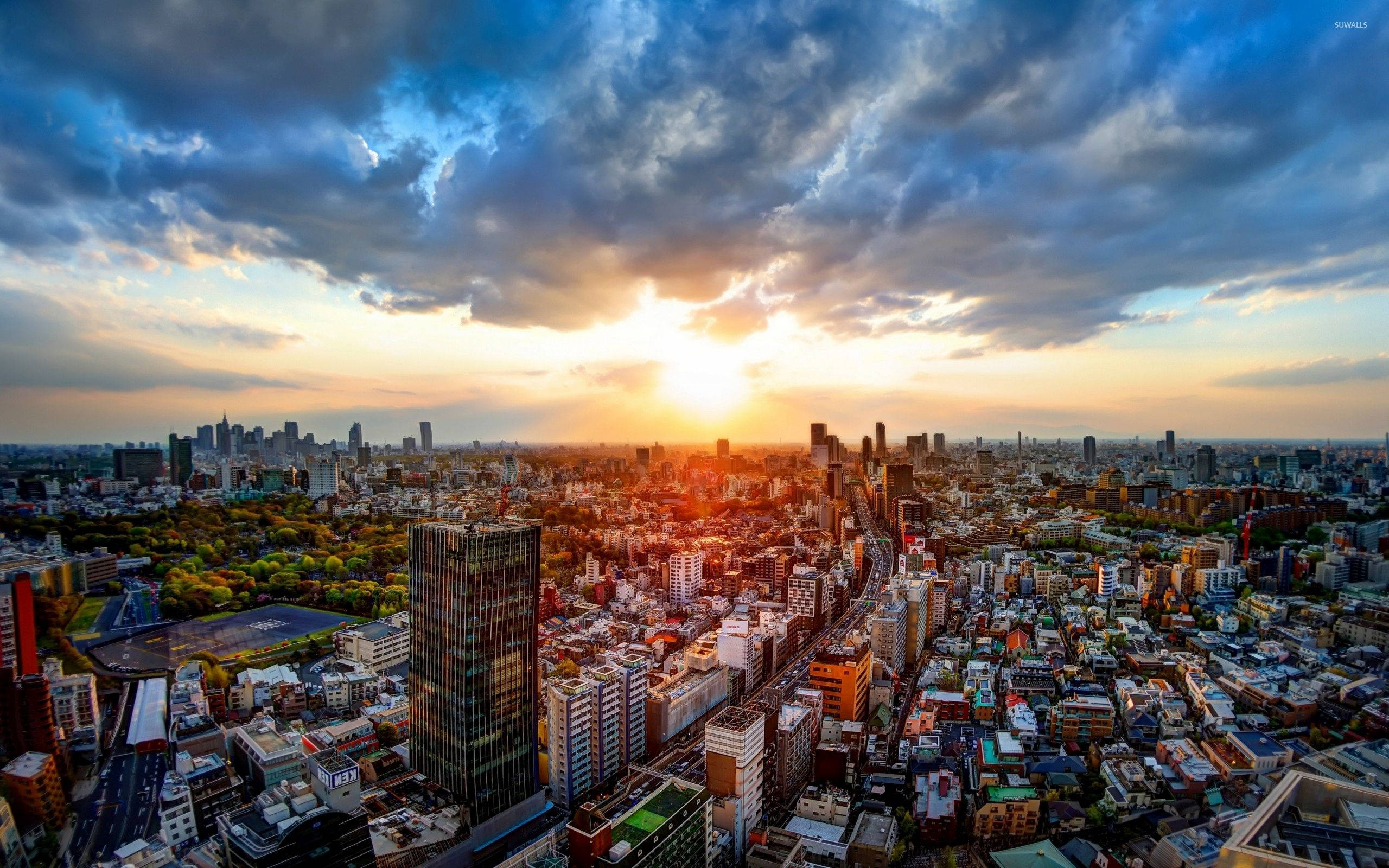 東京 桌布 壁紙 — 夕陽將廣闊的城市天際線沐浴在金色光芒中，戲劇性的雲層在密集的街區和綠色公園上空翻騰。