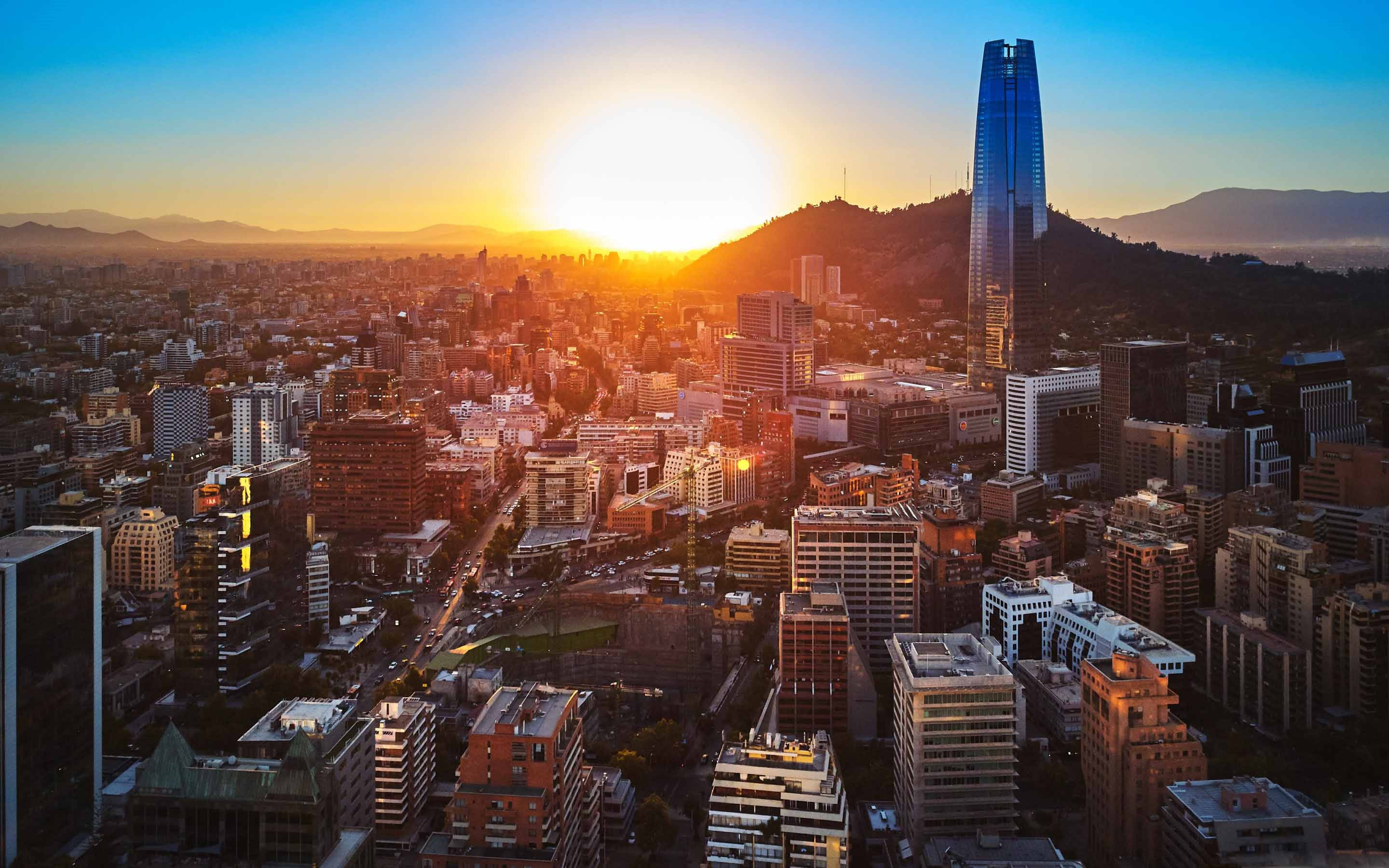 城市 桌布 壁紙 — 夕陽將廣闊的城市景觀沐浴在金色光芒中，一座高聳的摩天大樓矗立在密集的城市建築和遠山之上。