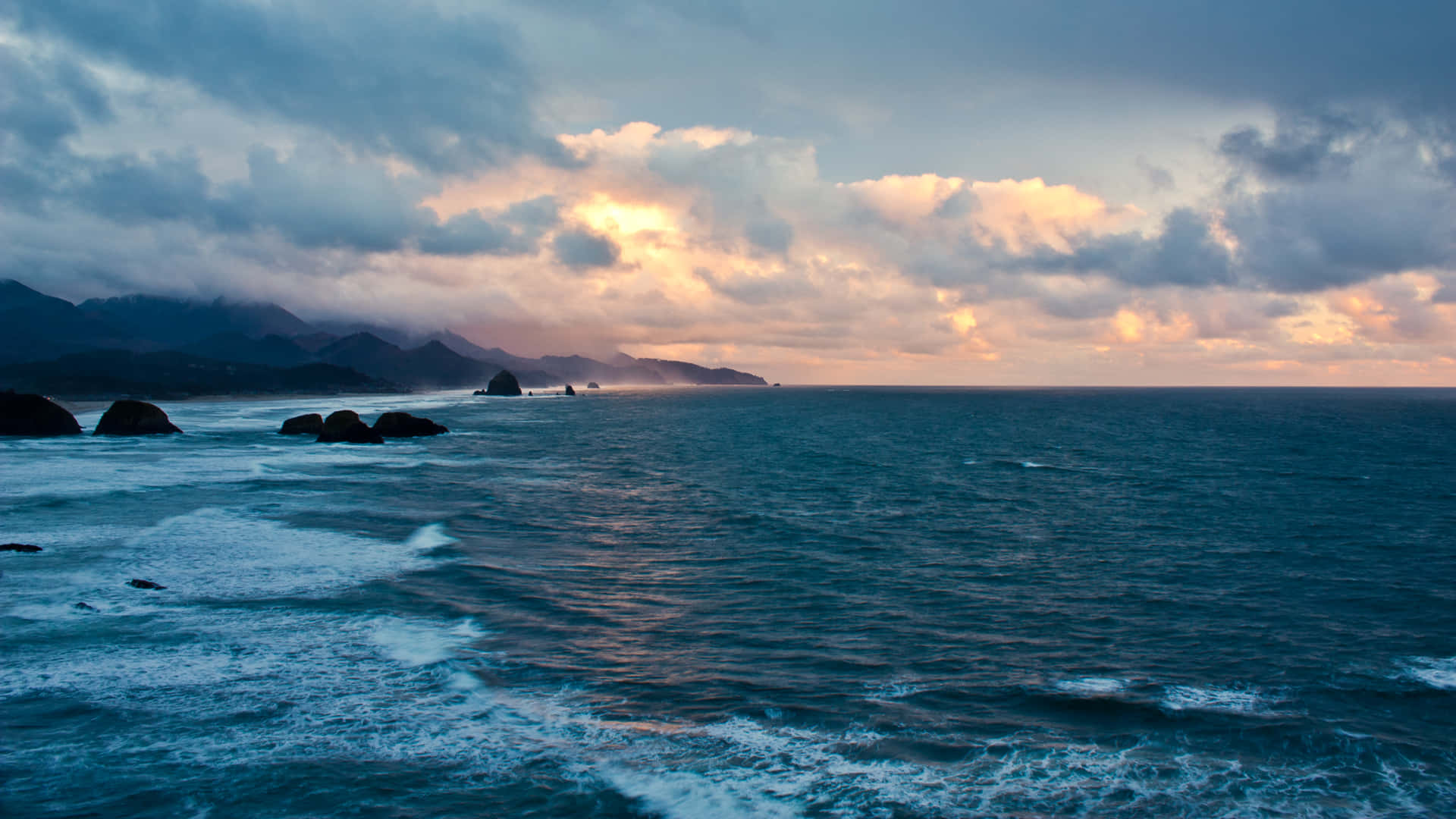 Pretty Ocean 桌布 壁紙 — 夕陽將暴風雲染成橙色，海浪拍打著崎嶇海岸線上的黑色岩石。