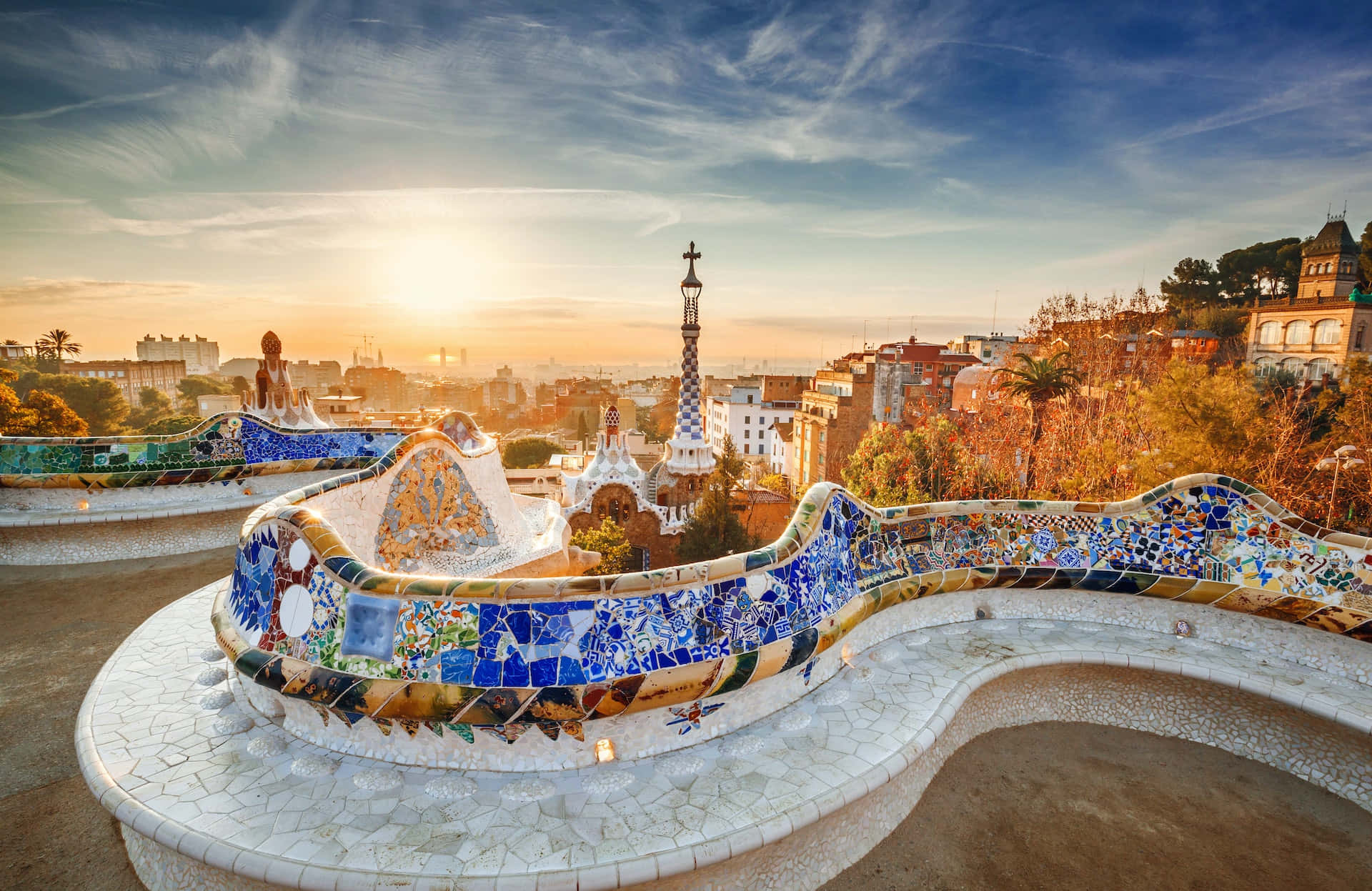 Barcelona 桌布 壁紙 — 夕陽將桂爾公園鋪滿馬賽克的蛇形長椅沐浴在溫暖的金色光芒中，背景是巴塞隆納的天際線。