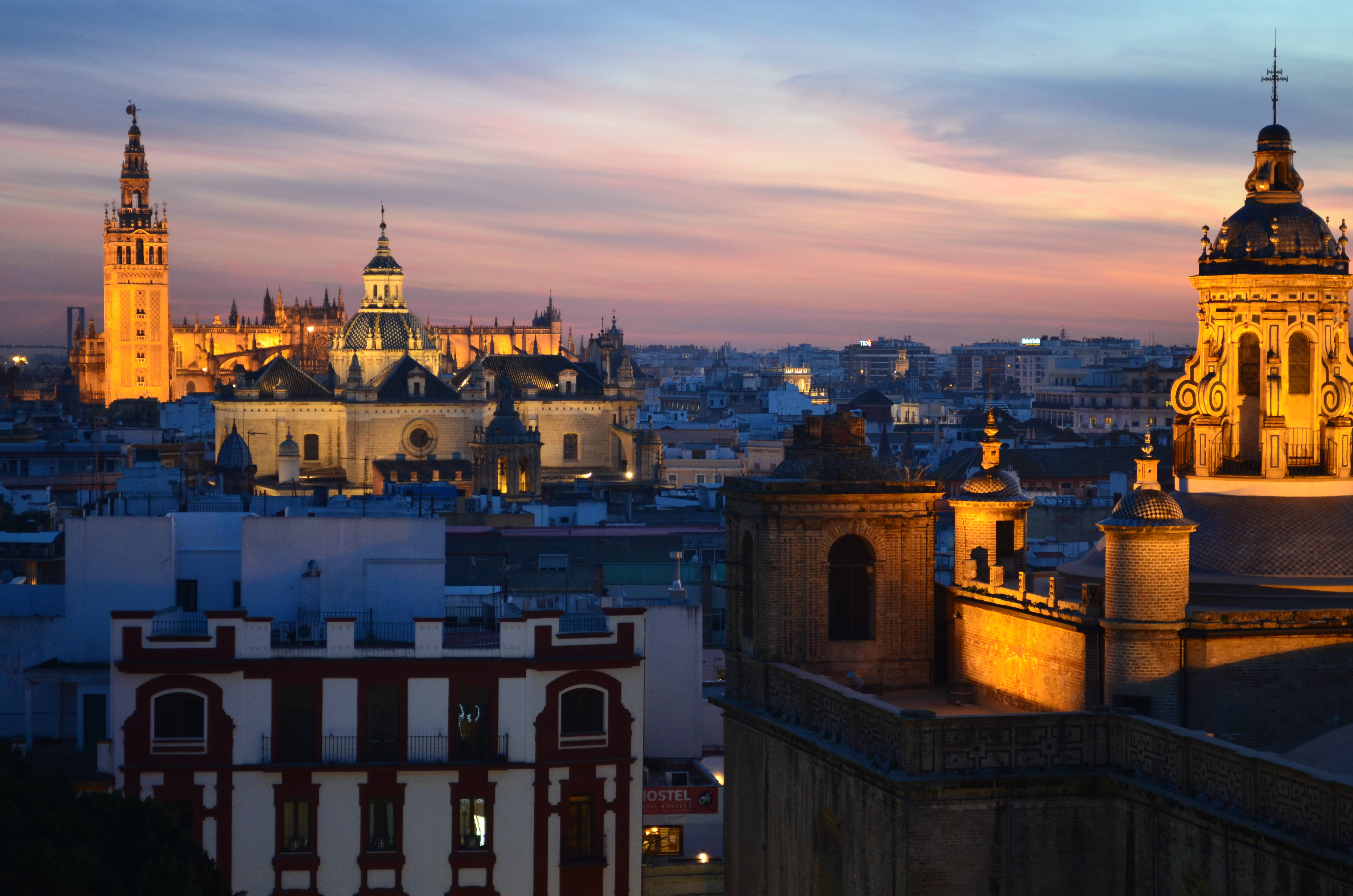 Seville 桌布 壁紙 — 夕陽將歷史悠久的塞維利亞大教堂和屋頂沐浴在溫暖的金色光芒中，映襯著柔和的粉紅色和藍色天空。