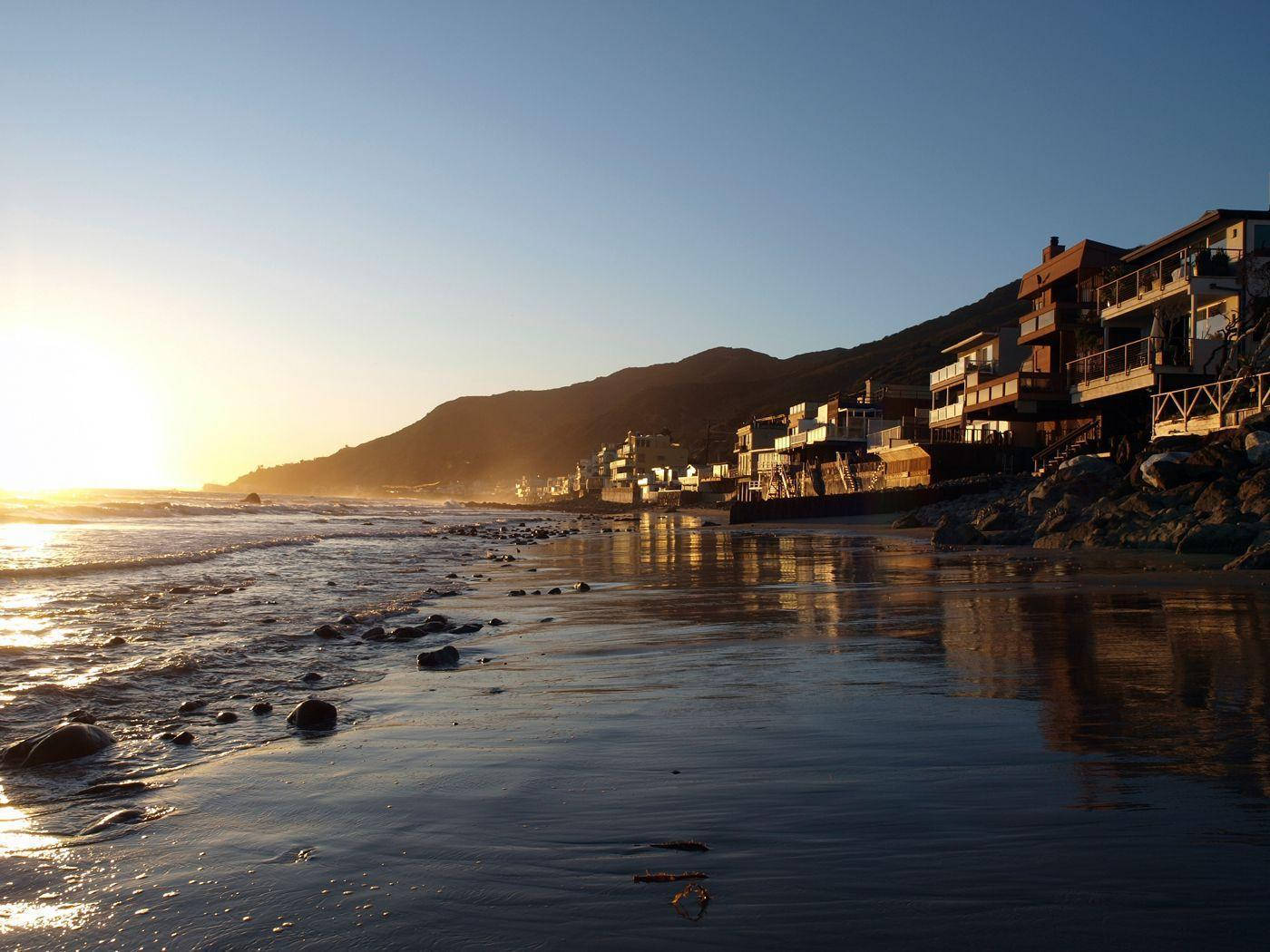 Malibu California 桌布 壁紙 — 夕陽將沿海小鎮沐浴在金色光芒中，海浪拍打著濕潤的沙灘，沙灘上散落著岩石。