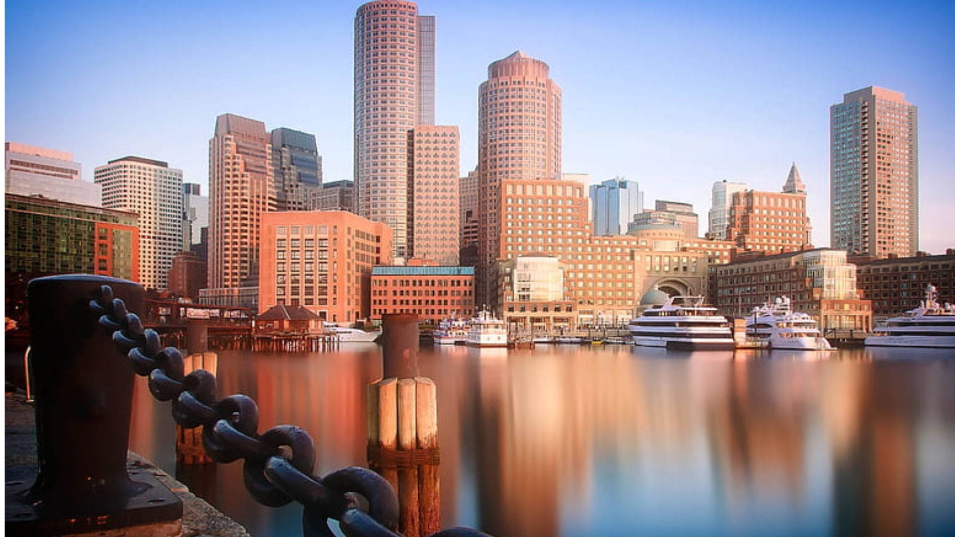 Massachusetts 桌布 壁紙 — 夕陽將波士頓港沐浴在溫暖的金色中，摩天大樓倒映在平靜的水面上，旁邊停泊著遊艇和沉重的鐵鏈圍欄。