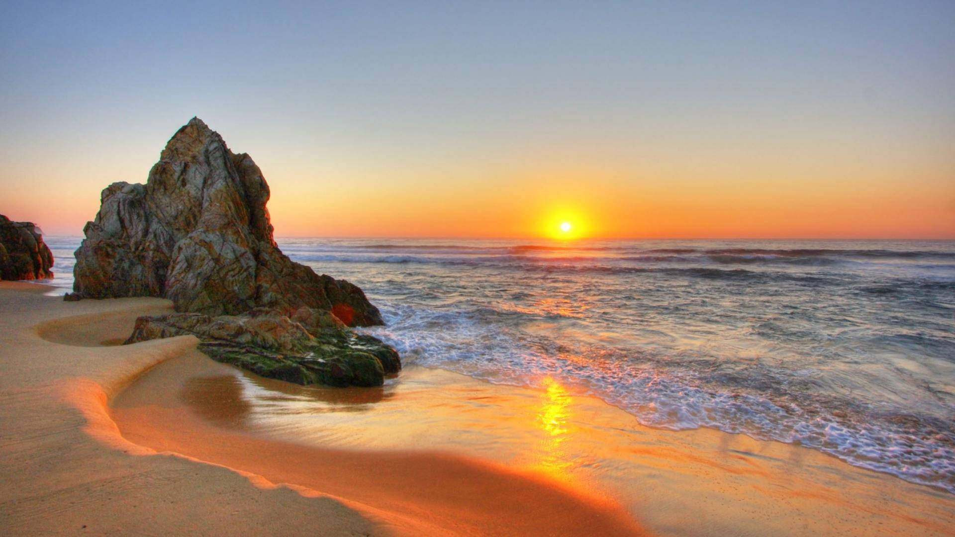 海灘日落 桌布 壁紙 — 夕陽將海洋和沙灘染成溫暖的橙色，海浪拍打著寧靜海灘上的一塊大岩石。