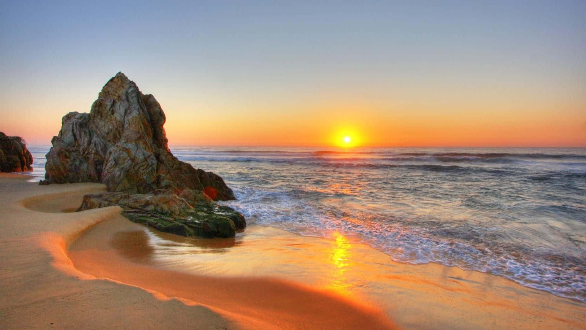 日落 桌布 壁紙 — 夕陽將海洋和沙灘染成溫暖的橙色色調，海浪輕輕拍打著一個巨大的岩石突出物。