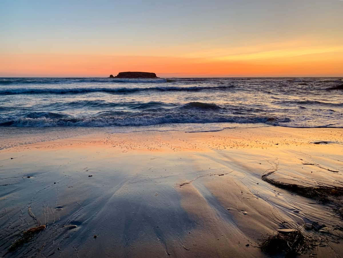 Oregon Coast 桌布 壁紙 — 夕陽將海洋和濕沙染成溫暖的橙色和粉紅色調，海浪向遠處的岩石島嶼翻滾。