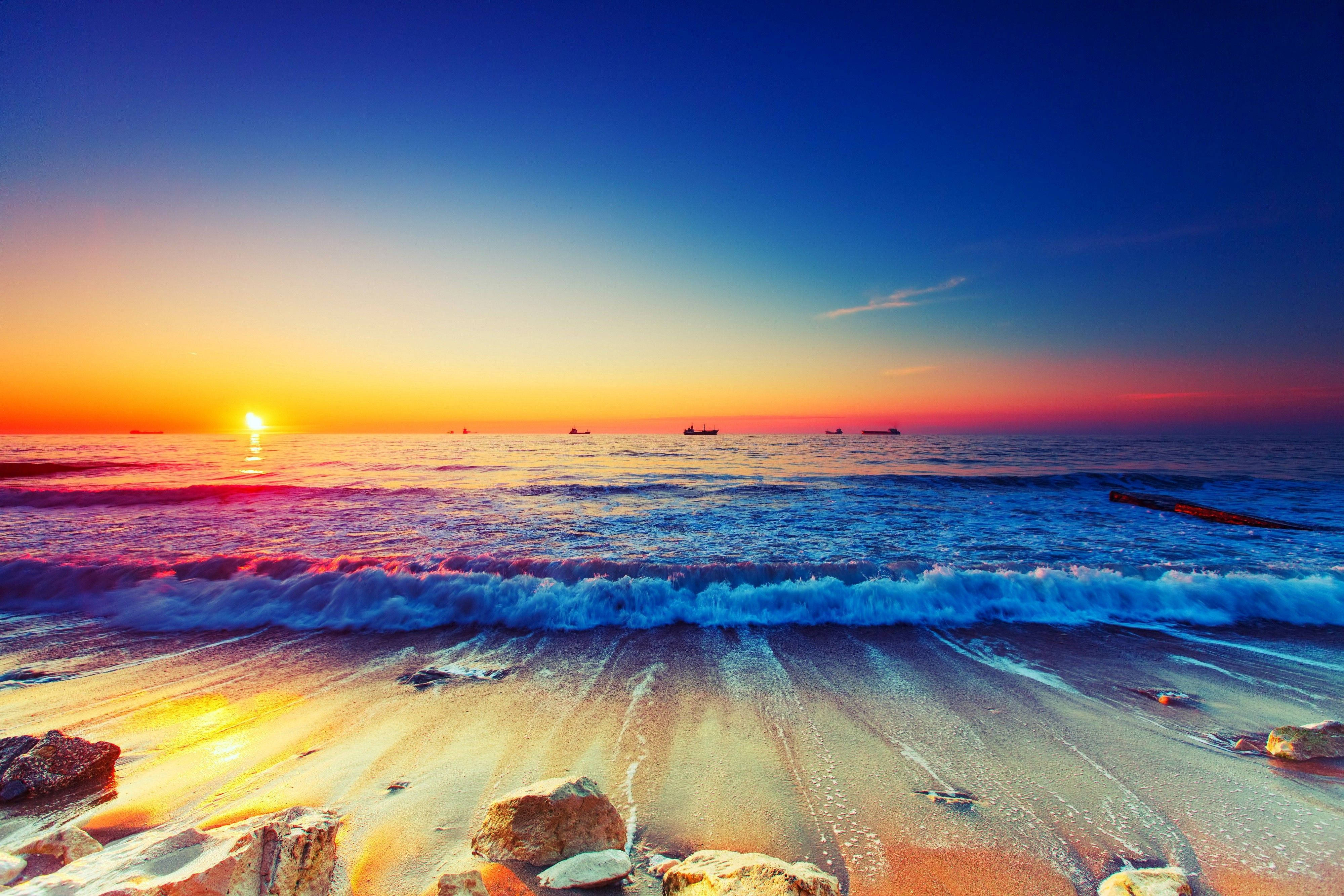海洋 桌布 壁紙 — 夕陽將海洋染成火橙色和深藍色，海浪拍打著佈滿岩石的沙灘。