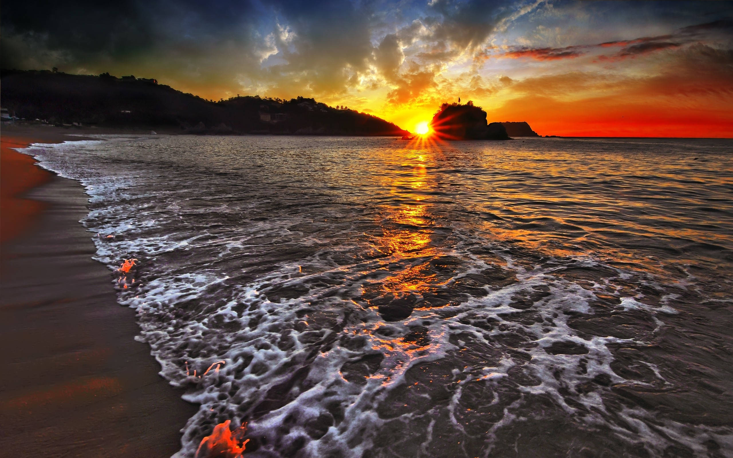 View 桌布 壁紙 — 夕陽將海洋染成火熱的橙色，海浪在戲劇性的雲層下拍打著沙灘。