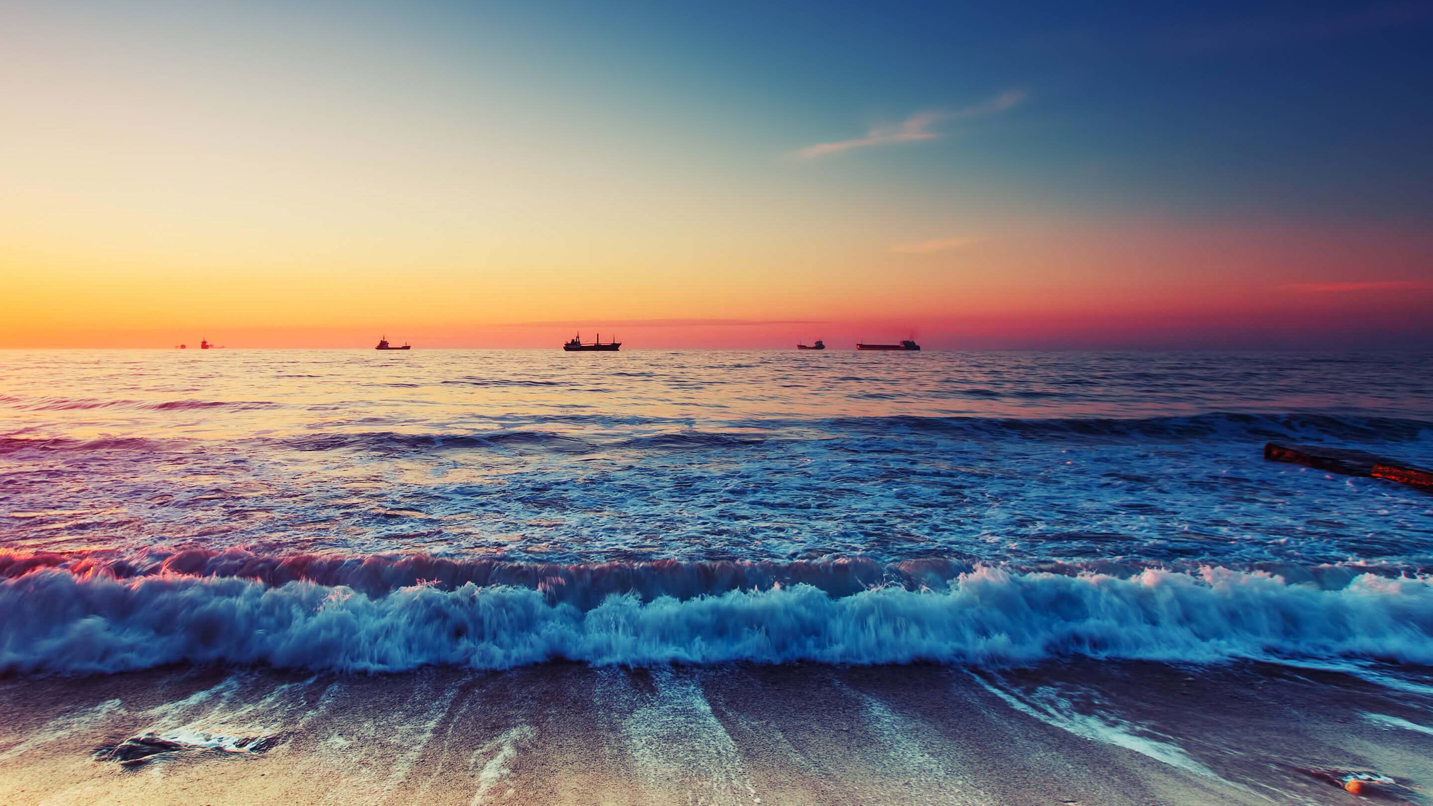 海洋 桌布 壁紙 — 夕陽將海洋染成火熱的橙色和深藍色，海浪拍打沙灘，遠處的船隻在地平線上形成剪影。