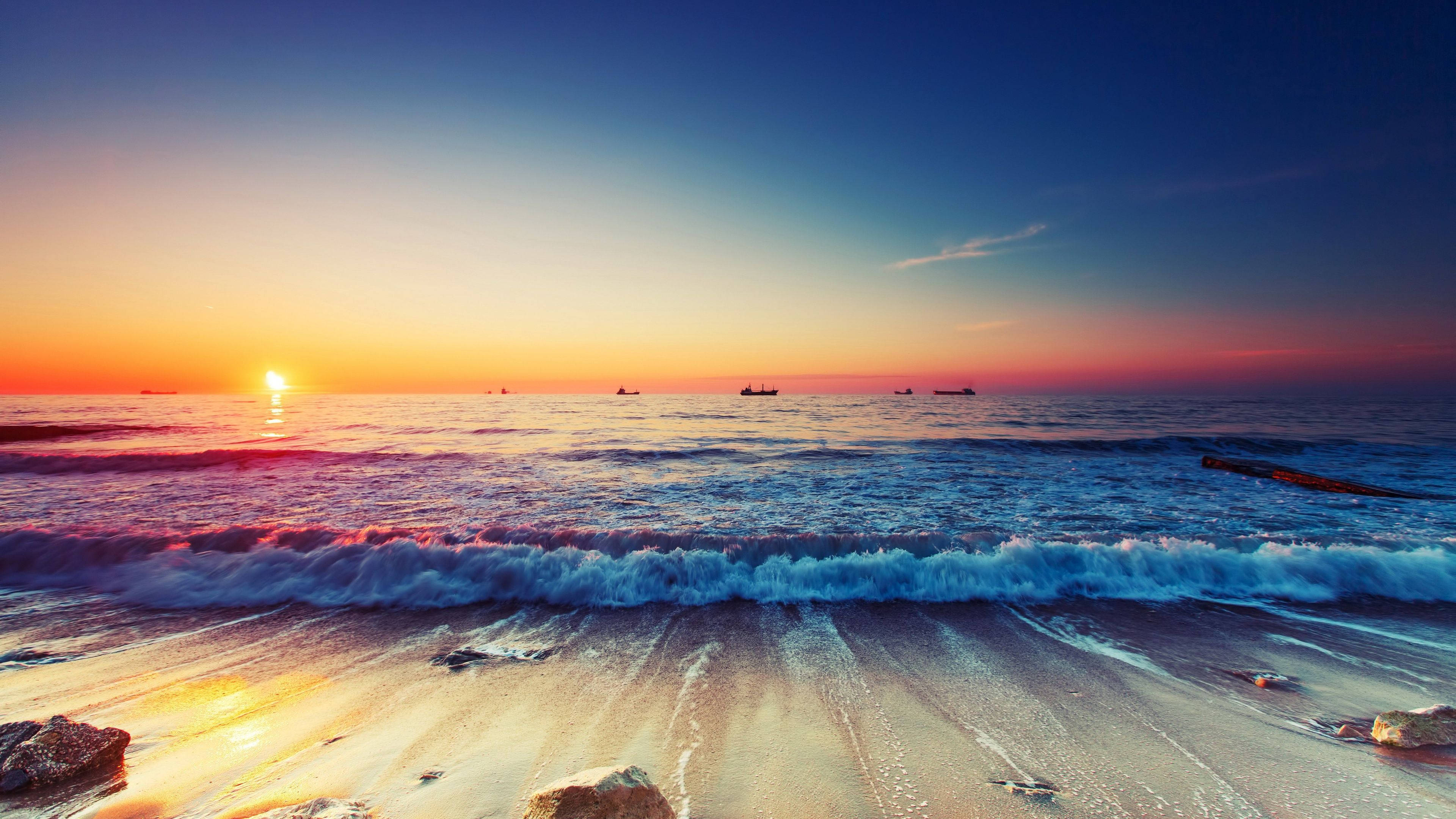 極高畫質 桌布 壁紙 — 夕陽將海洋染成火熱的橙色和深藍色，海浪拍打沙灘，遠處的船隻在地平線上形成剪影。