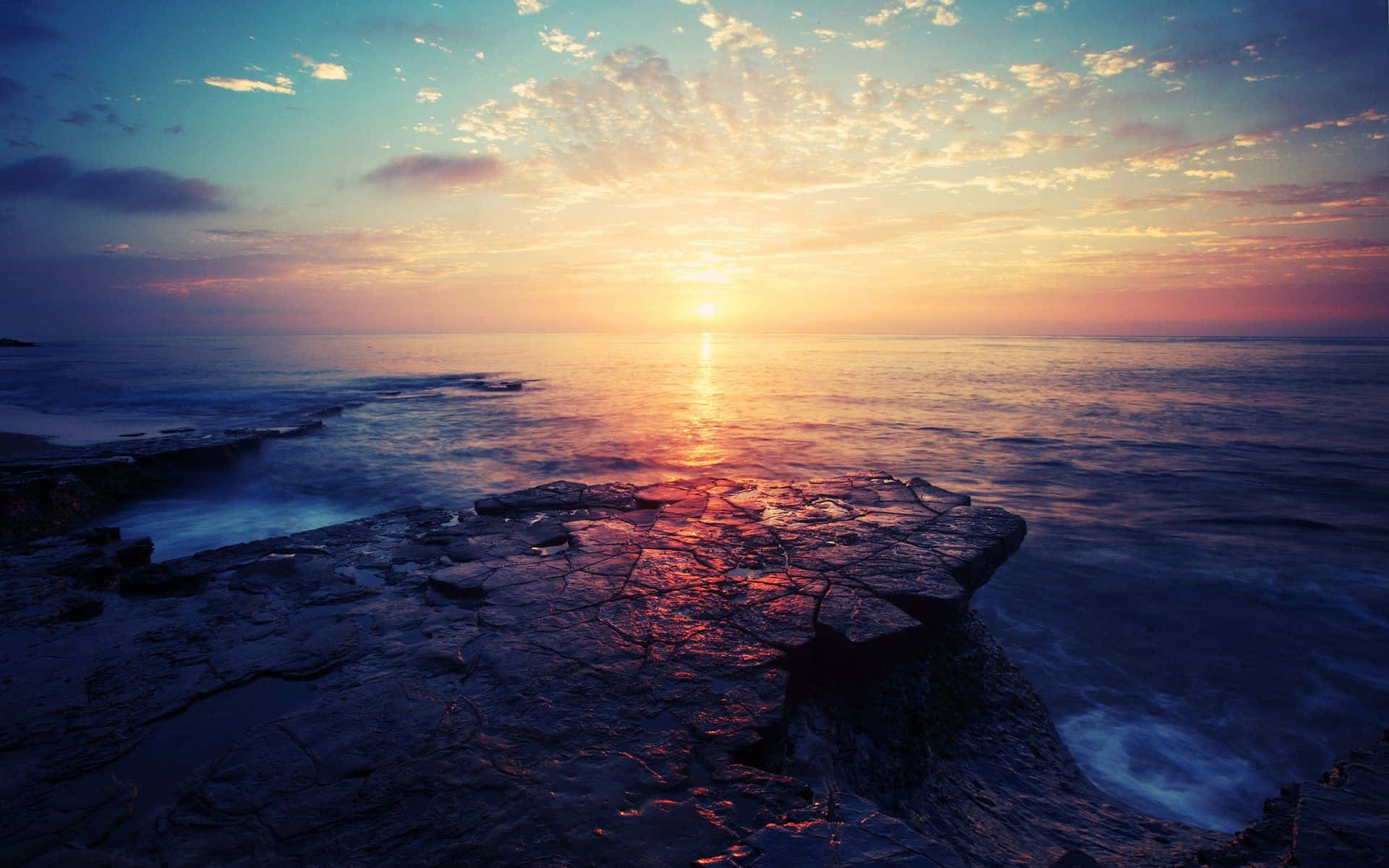 日出 桌布 壁紙 — 夕陽將海洋染成火熱的橙色和紫色，海浪拍打著黑暗的岩石海岸。