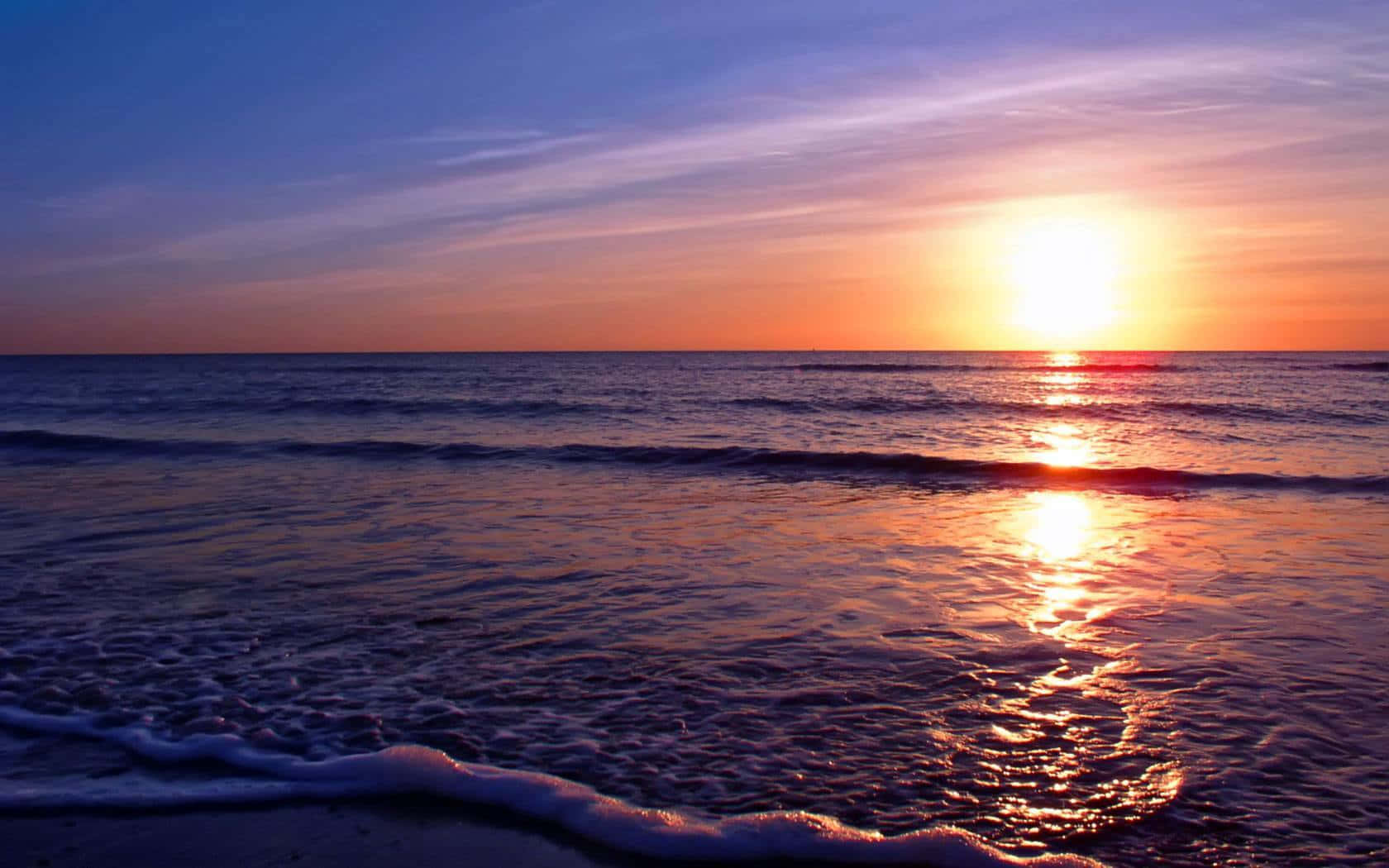 桌布 壁紙 — 夕陽將海洋染成火熱的橙色和紫色，溫柔的海浪在柔和的暮色天空下輕拍著海岸。