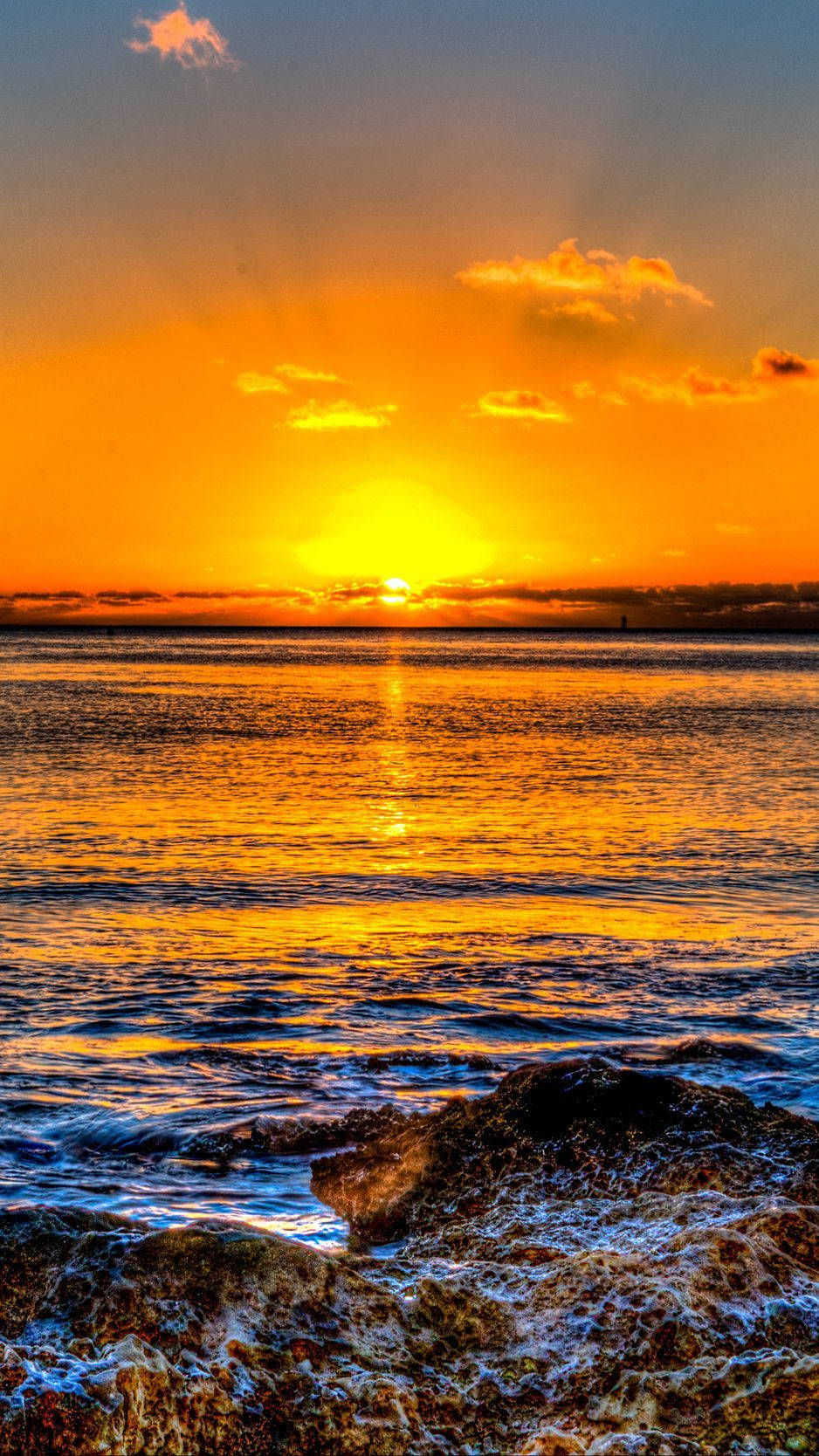 Sunset iPhone 桌布 壁紙 — 夕陽將海洋染成火熱的橙色和金色，海浪拍打前景的黑色岩石。
