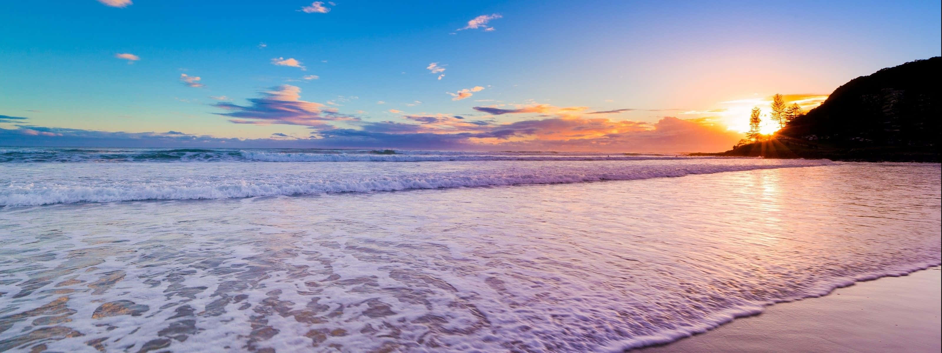 Beach Dual Monitor 桌布 壁紙 — 夕陽將海洋染成金色，海浪在充滿活力的天空下拍打沙灘。