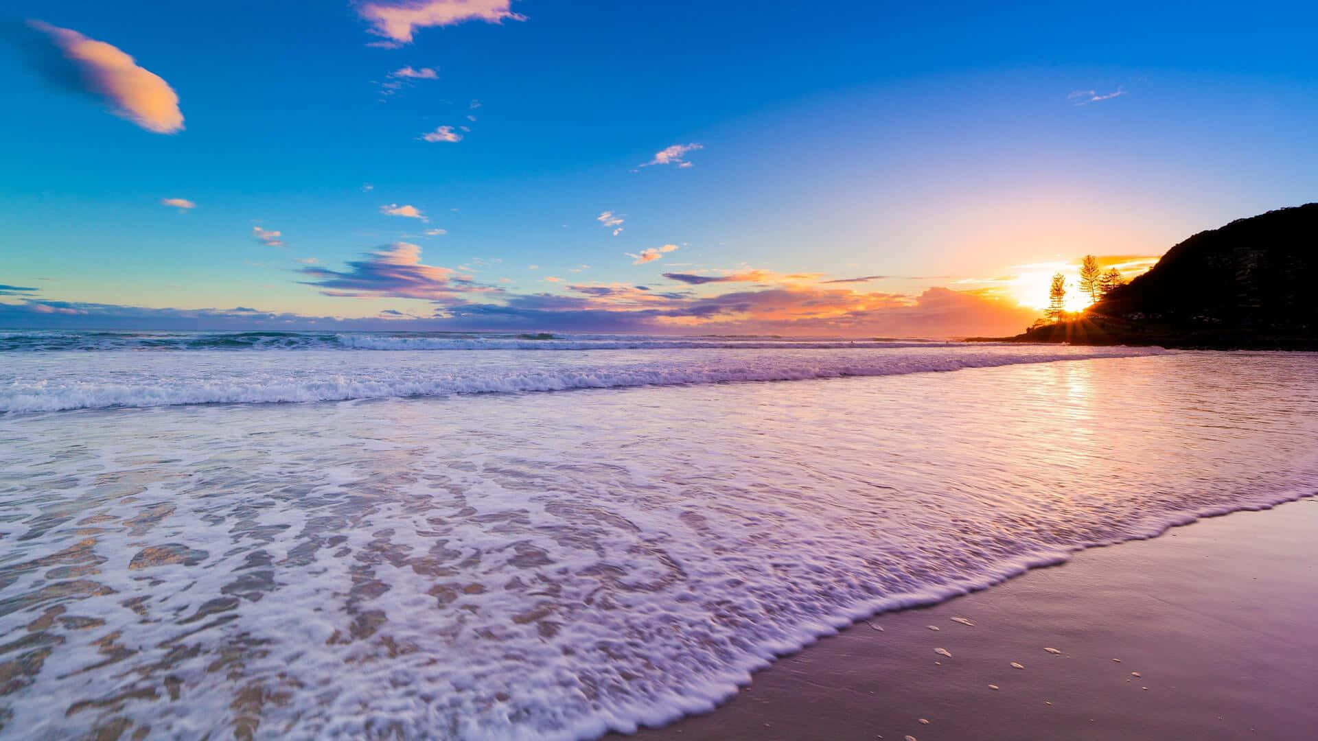 海灘日落 桌布 壁紙 — 夕陽將海浪和天空染成溫暖的金色和紫色，溫柔的浪花拍打著沙灘。