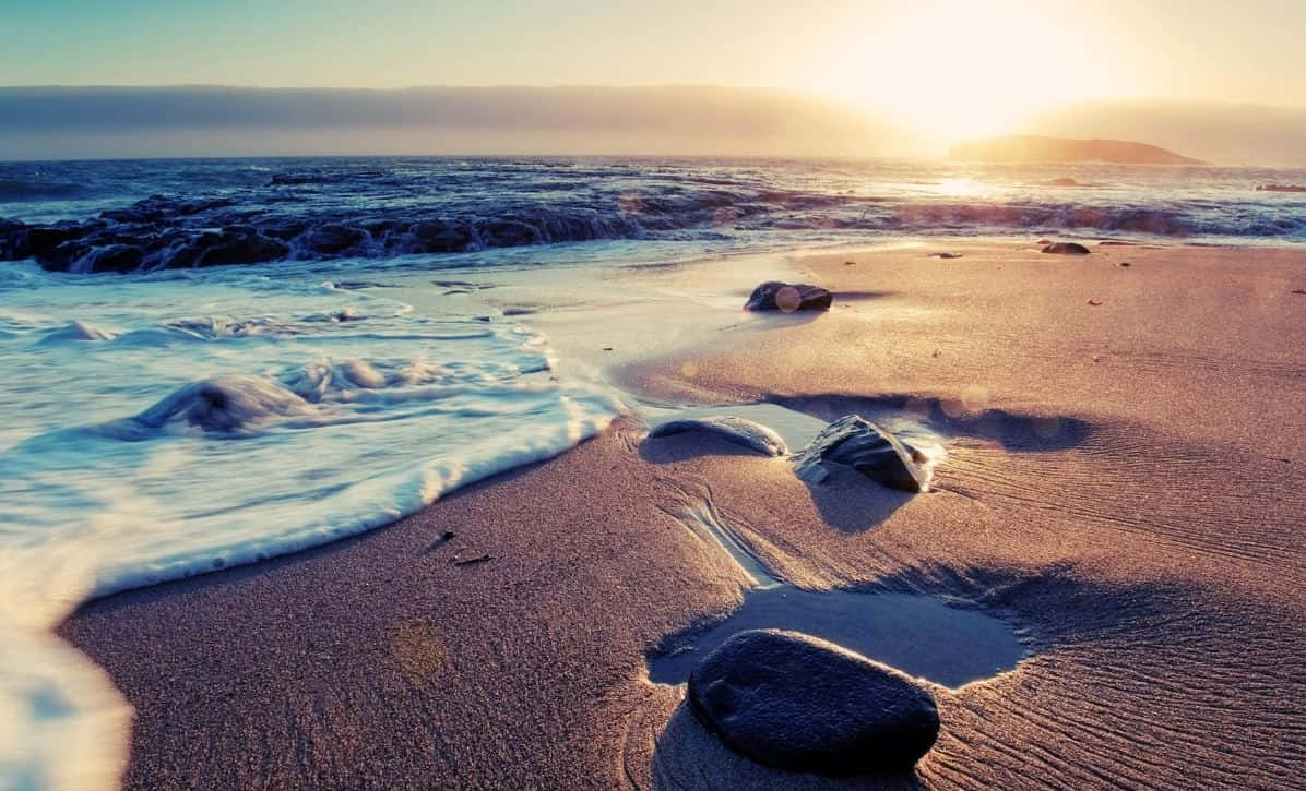 Seashore 桌布 壁紙 — 夕陽將海浪和沙灘染成溫暖的金色和清涼的藍色，溫柔的波浪拍打著散落的岩石。