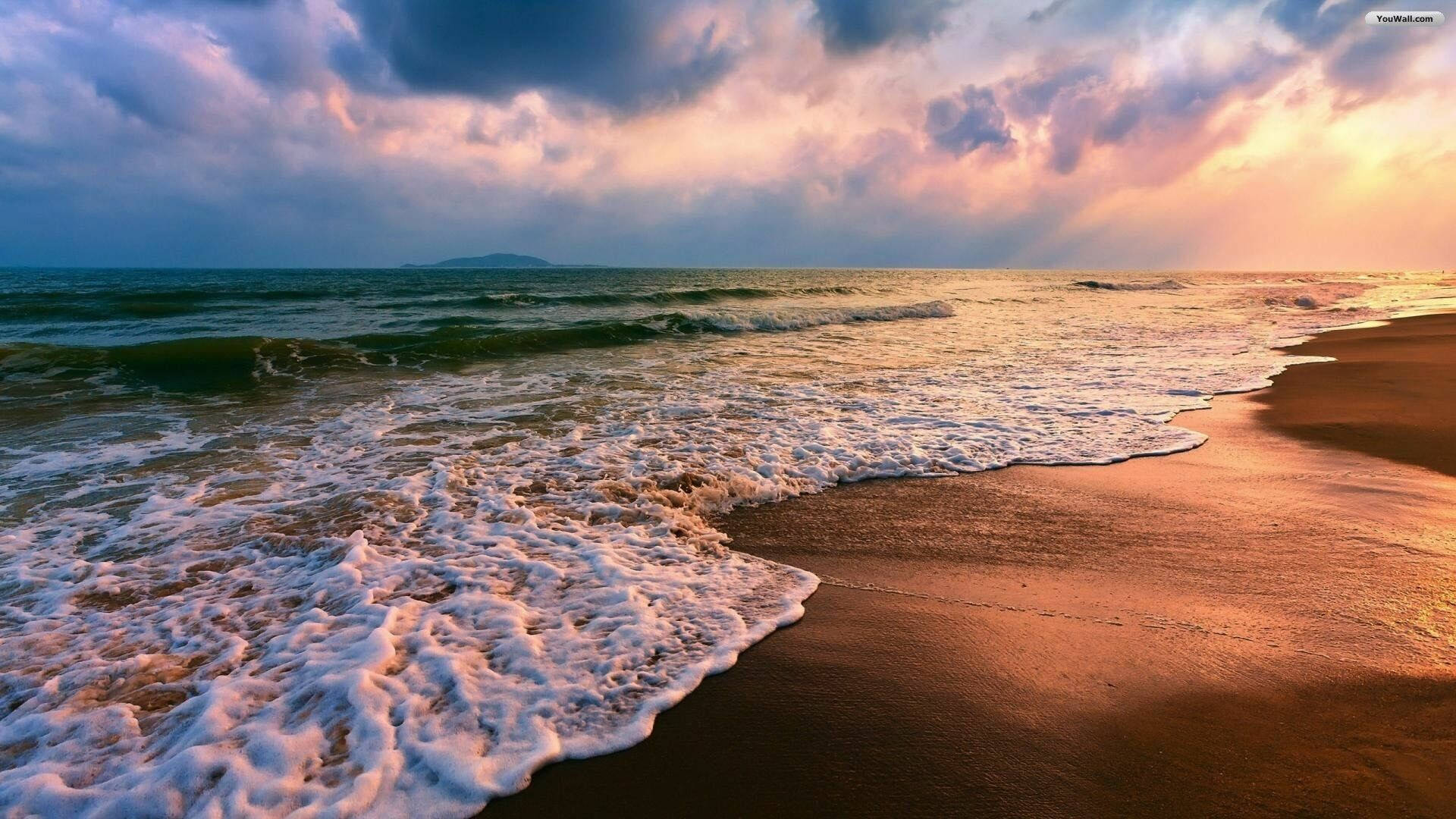 Beach Desktop 桌布 壁紙 — 夕陽將海浪和沙灘染成溫暖的金色和紫色，天空佈滿戲劇性的雲彩。