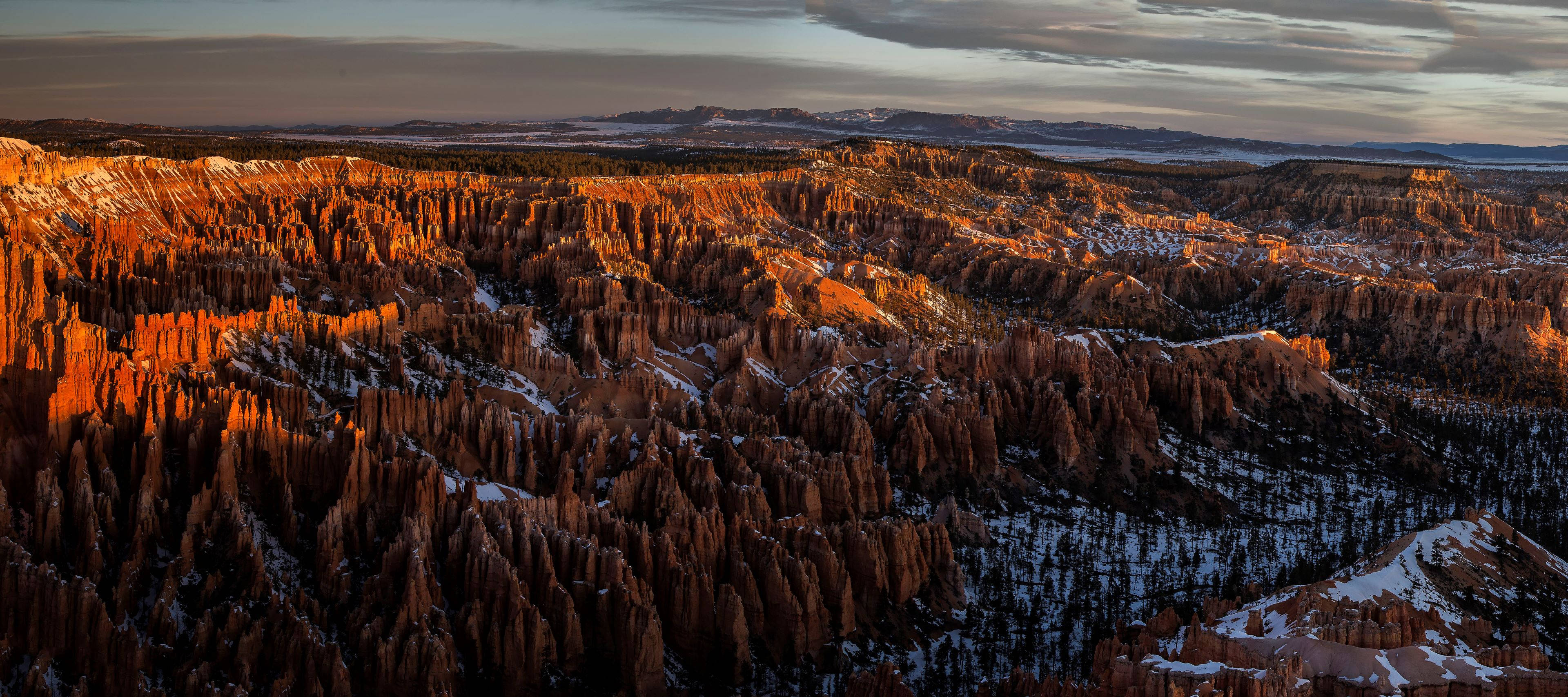 峽谷 桌布 壁紙 — 夕陽將灑滿白雪的岩柱（hoodoos）沐浴在溫暖的橙色光芒中，橫跨廣闊的峽谷景觀。