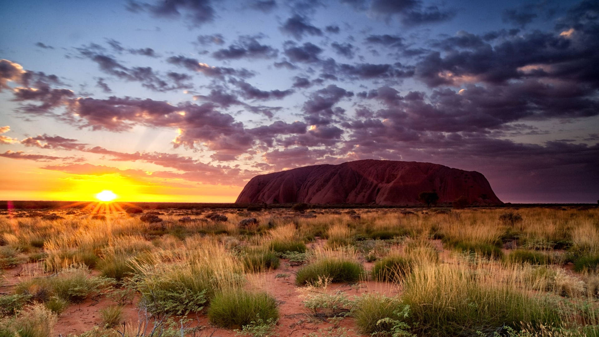 Outback 桌布 壁紙 — 夕陽將烏魯魯沐浴在溫暖的金色中，雲彩在乾燥的澳洲內陸上空呈現紫色和橙色條紋。