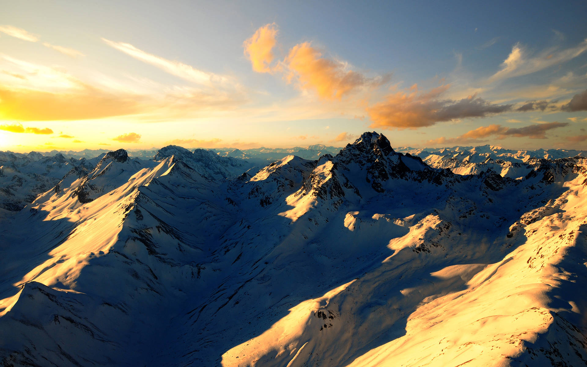高清 桌布 壁紙 — 夕陽將積雪的山峰沐浴在金色光芒中，映襯著柔和藍天與縷縷薄雲。