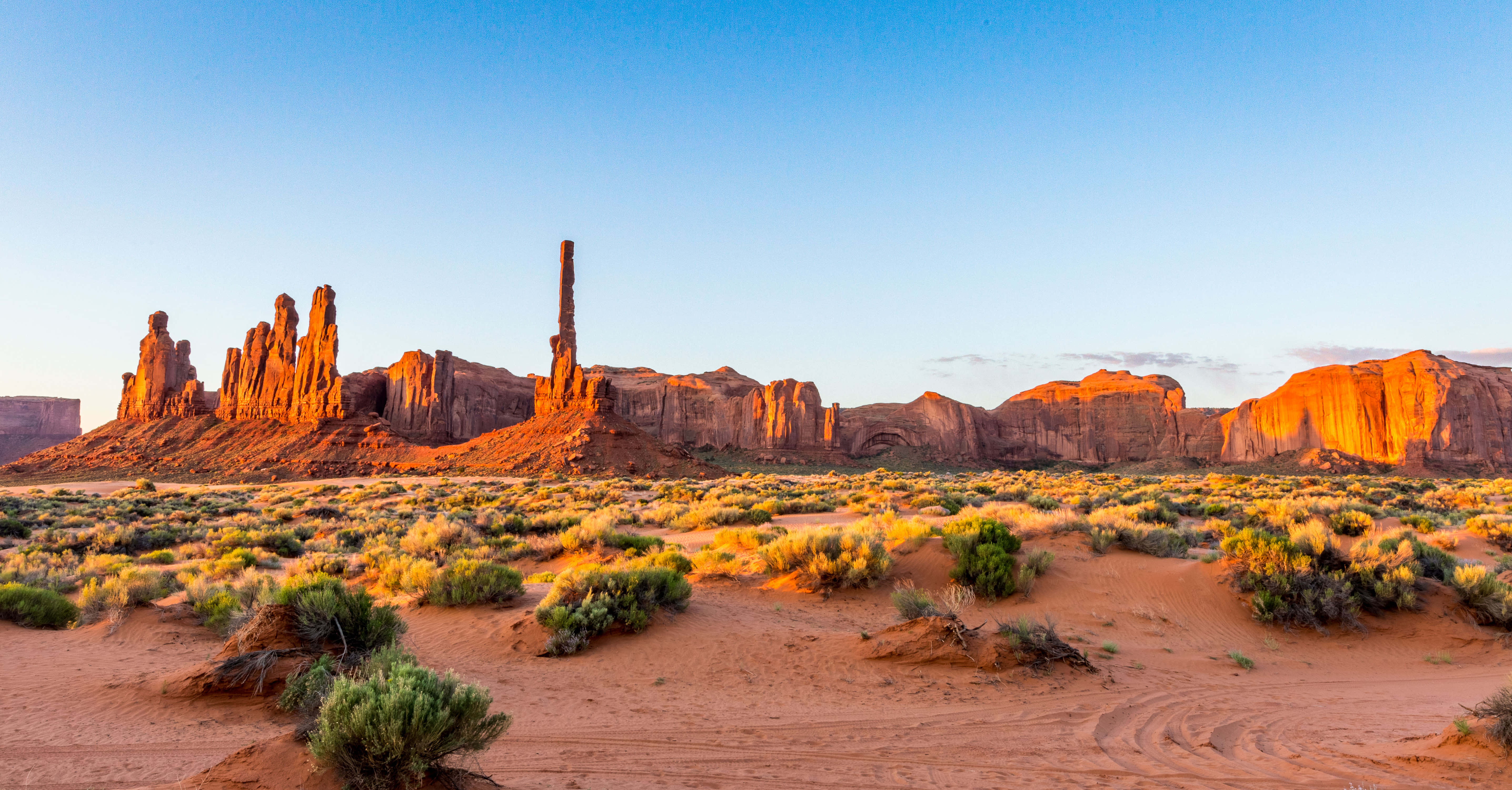 Arizona 桌布 壁紙 — 夕陽將紀念碑谷標誌性的紅色砂岩但丁沐浴在溫暖的金色光芒中，映襯著清澈的藍天。