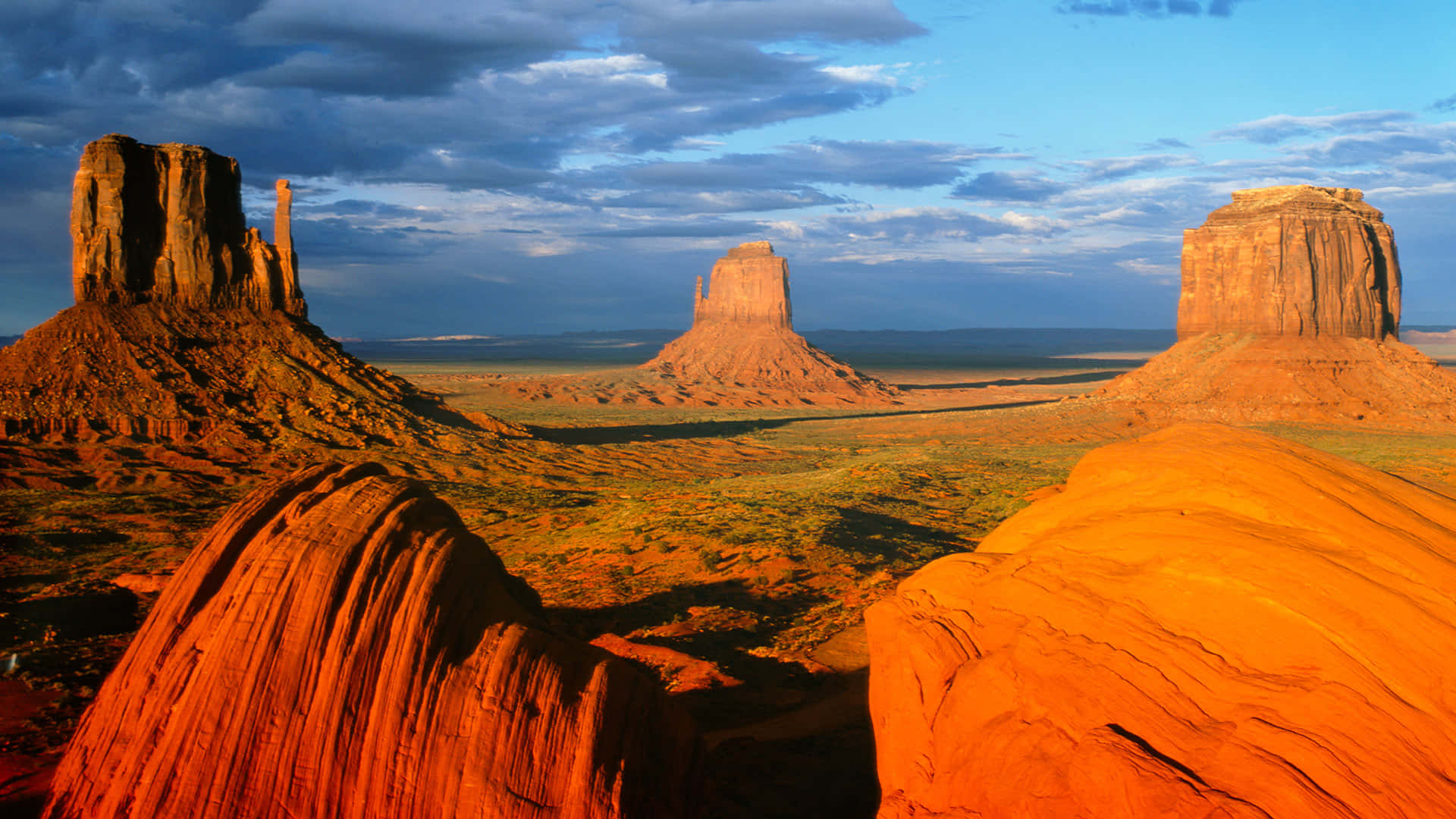 Arizona 桌布 壁紙 — 夕陽將紀念碑谷標誌性的紅色砂岩但丁山沐浴在溫暖的金色光芒中，映襯著帶有散雲的戲劇性藍天。