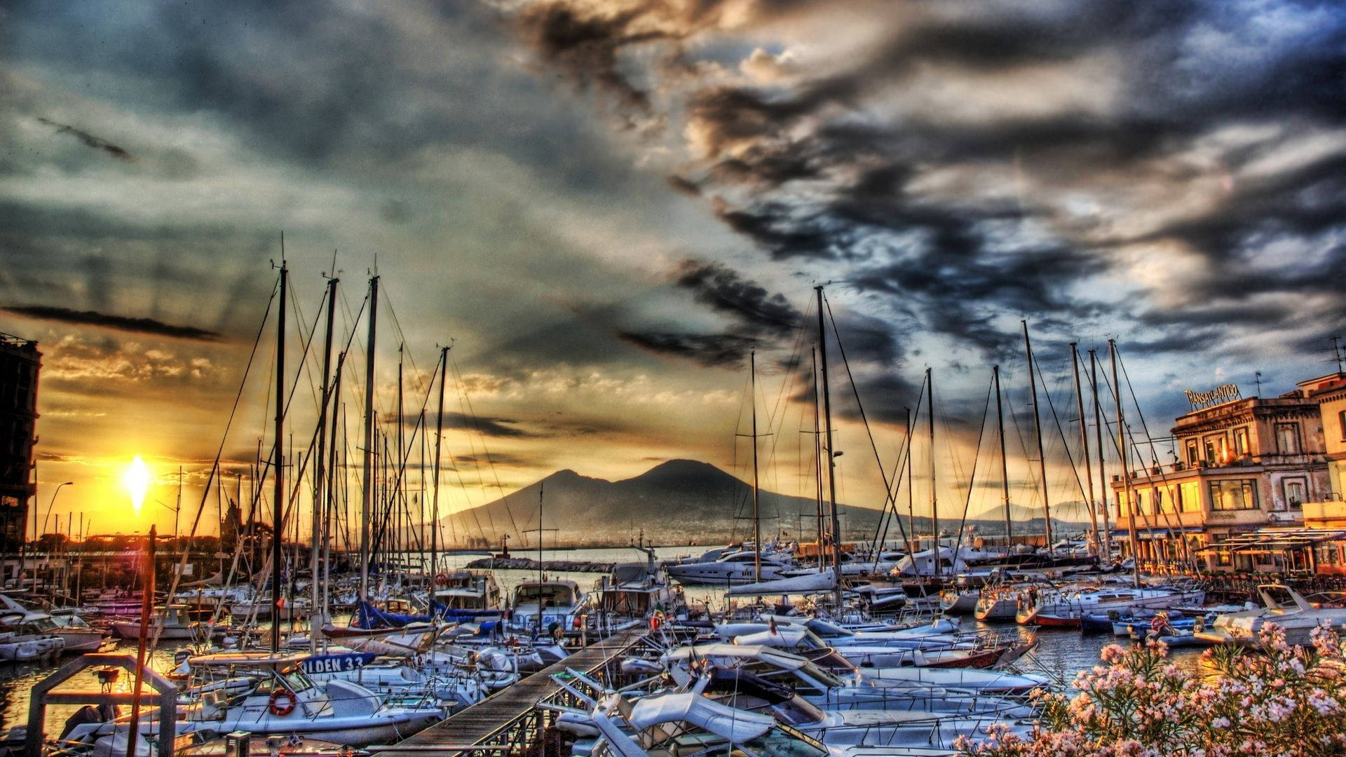 Naples 桌布 壁紙 — 夕陽將繁忙的碼頭沐浴在金色光芒中，帆船點綴水面，遠處維蘇威火山巍然聳立。
