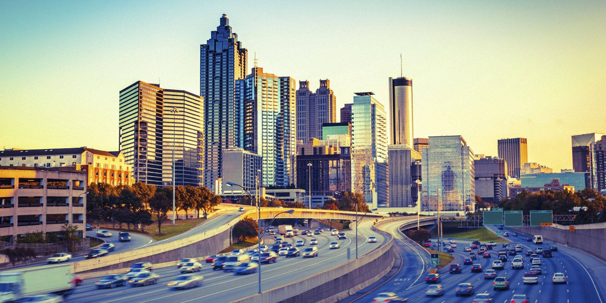 Atlanta 桌布 壁紙 — 夕陽將繁華的城市景觀沐浴在溫暖的金色光芒中，車流沿著彎曲的高速公路在高聳的玻璃摩天大樓下流動。