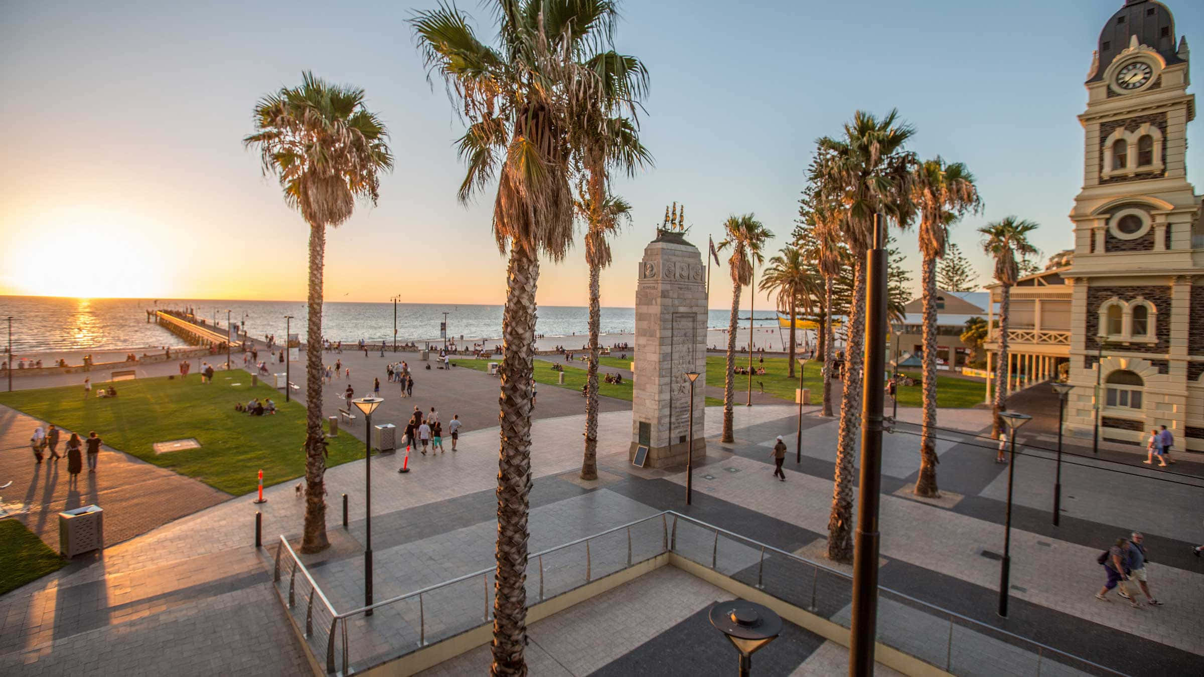 Adelaide 桌布 壁紙 — 夕陽將金色光芒灑在海岸廣場上，棕櫚樹在歷史悠久的鐘樓和海濱長廊旁搖曳。