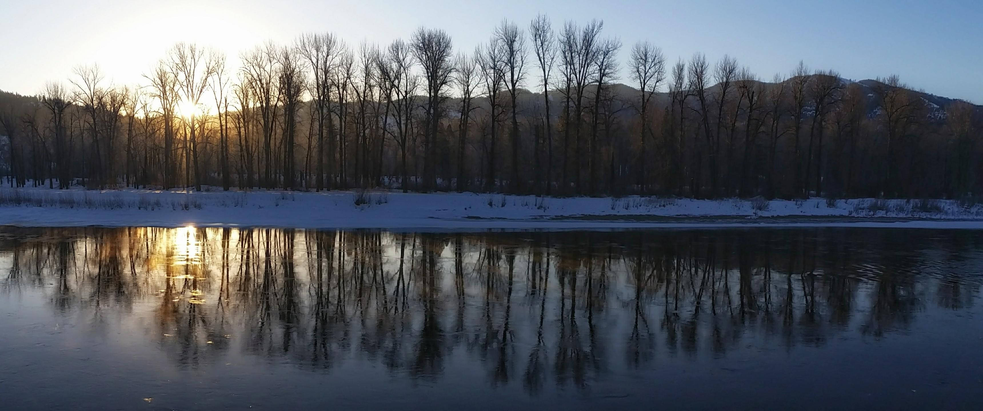 冬天 桌布 壁紙 — 夕陽將金色光芒灑在積雪覆蓋的樹木上，倒映在平靜的河流中，遠處有山巒。