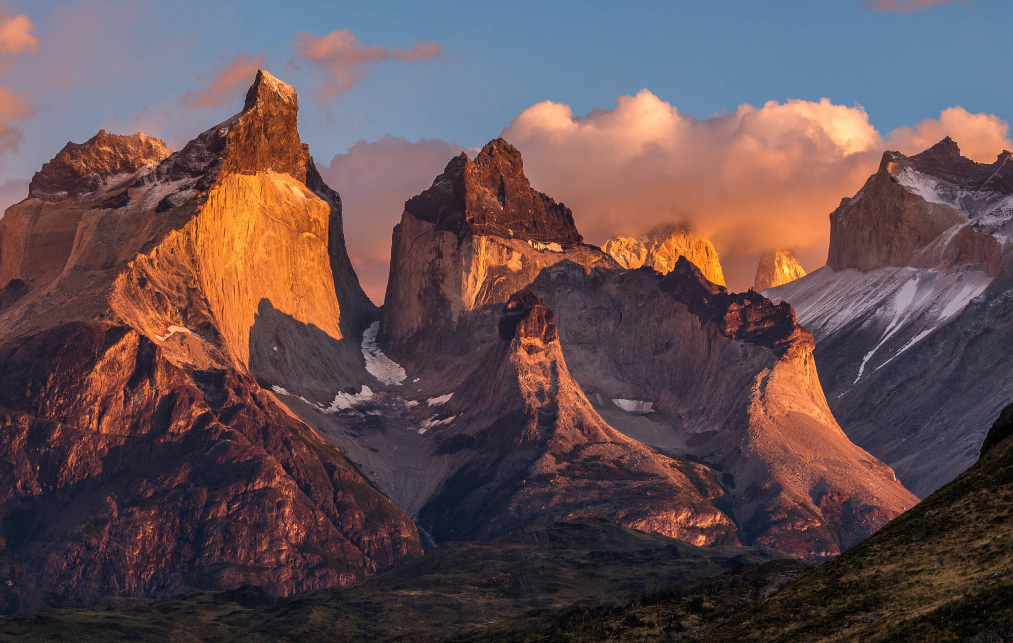 Patagonia 桌布 壁紙 — 夕陽將鋸齒狀的山峰沐浴在金色光芒中，雲朵在積雪的懸崖上方閃耀著粉紅色和橙色。
