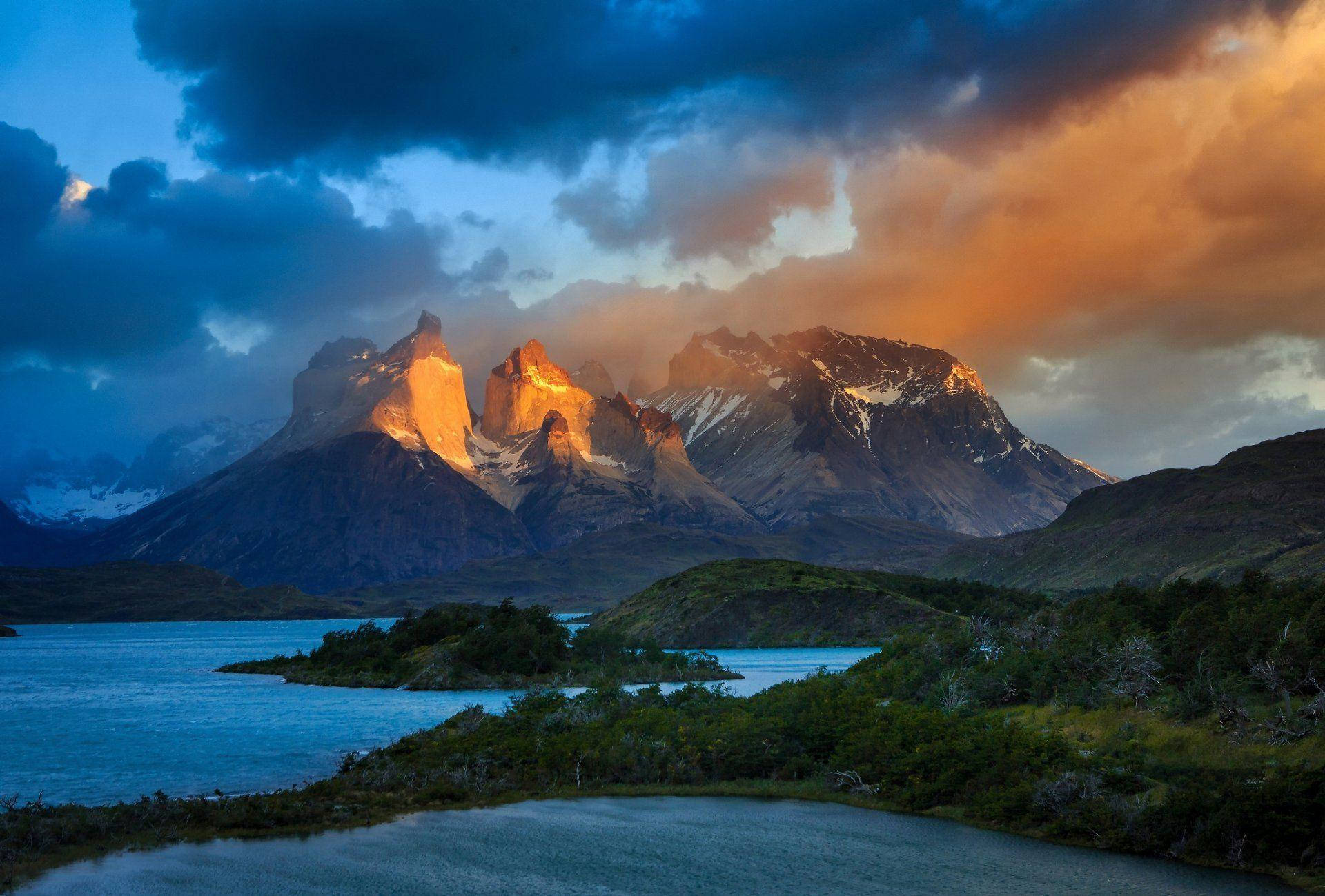 South America 桌布 壁紙 — 夕陽將鋸齒狀的山峰沐浴在金色光芒中，映照在平靜的藍色湖泊上，四周環繞著綠色山丘。