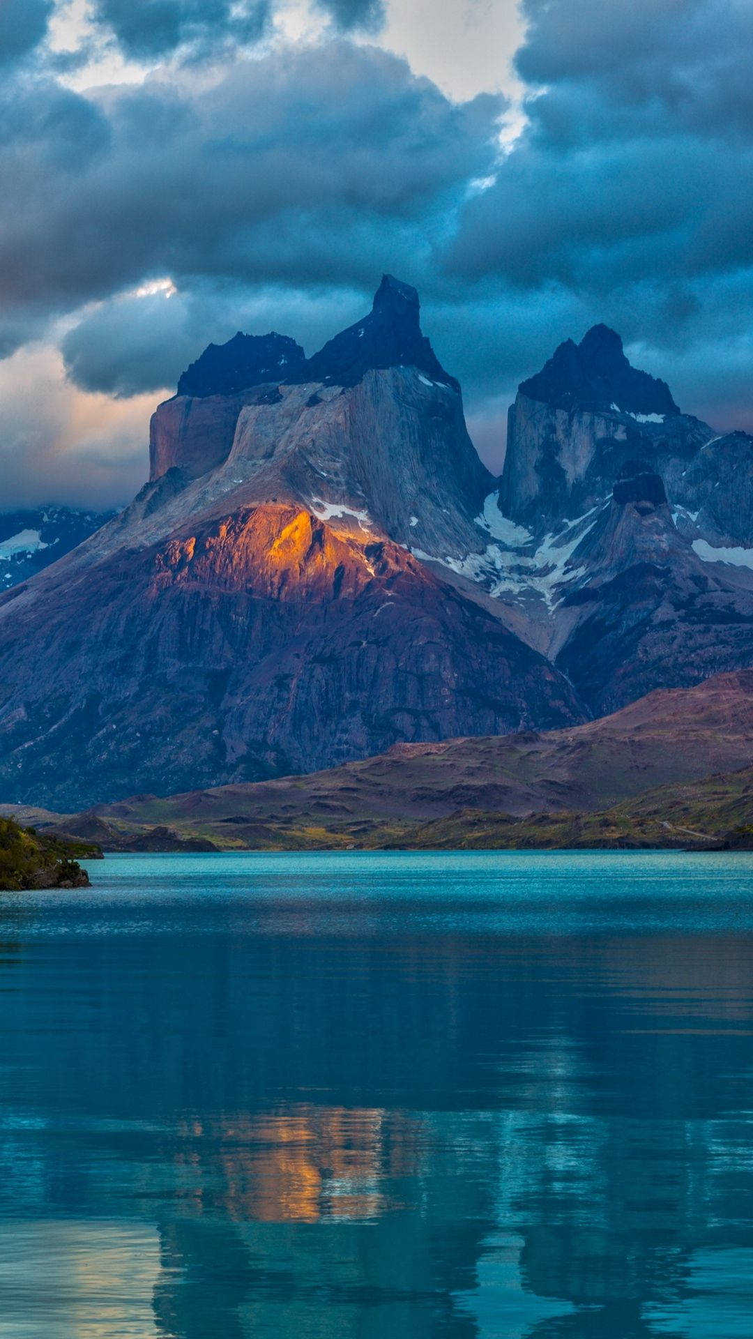 Patagonia 桌布 壁紙 — 夕陽將鋸齒狀的山峰沐浴在金色的光芒中，反射在平靜的碧綠湖面上，天空佈滿戲劇性的雲彩。