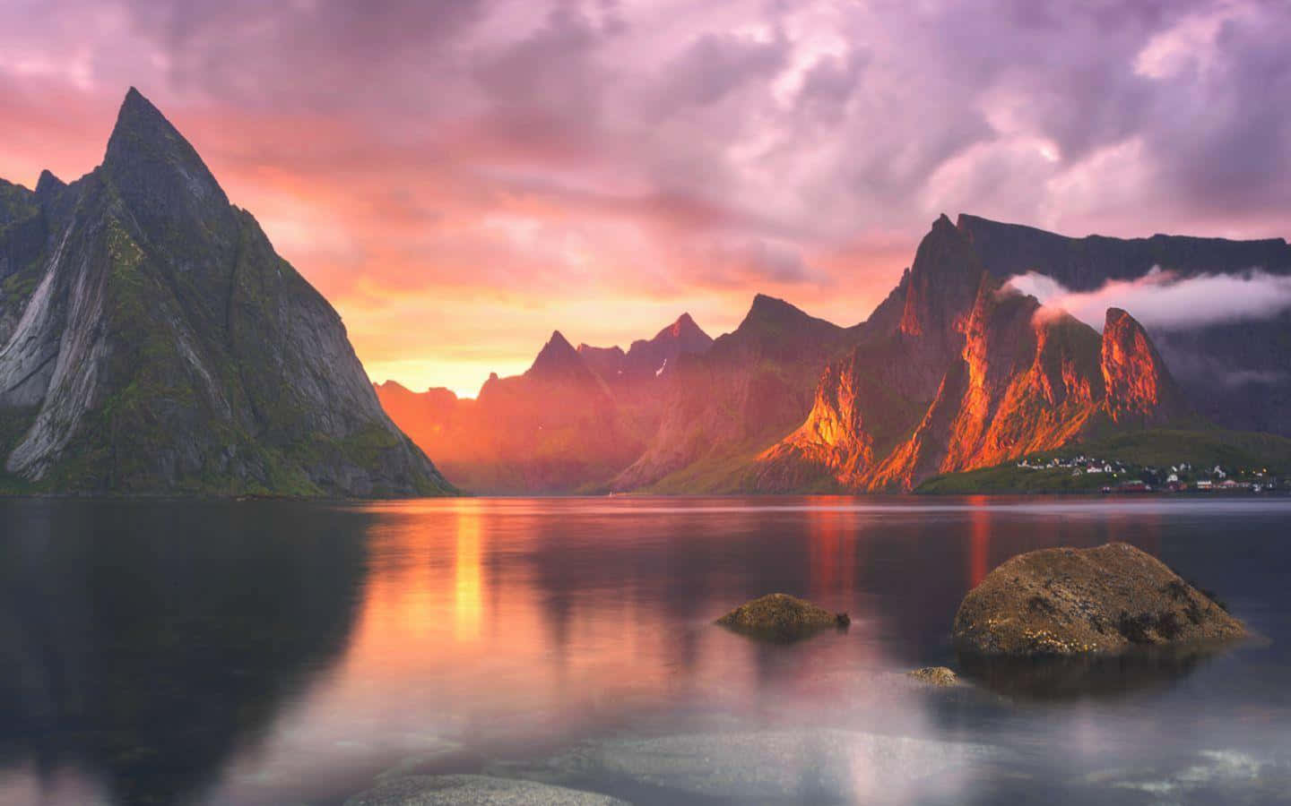 macOS 桌布 壁紙 — 夕陽將鋸齒狀的山脈和寧靜的水面染成火熱的橙色和紫色色調，前景有漂浮的岩石。