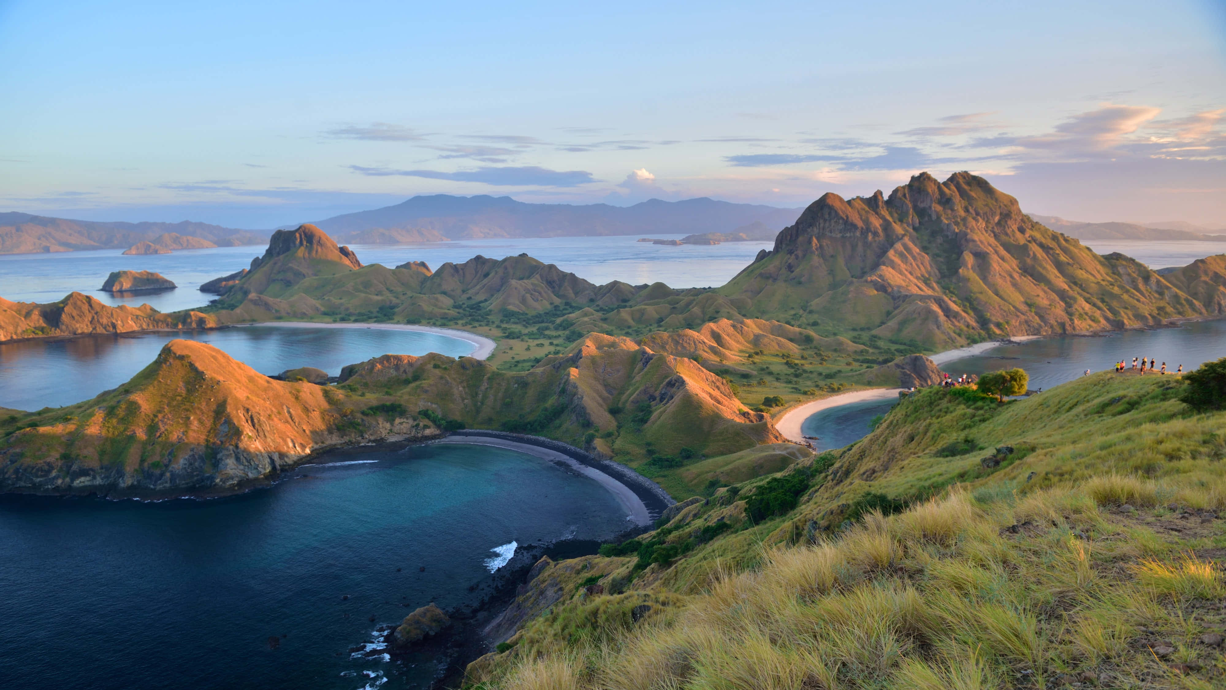 風景 桌布 壁紙 — 夕陽將鋸齒狀的火山島和碧綠的海灣沐浴在溫暖的金色光芒中，遠處的山脈在柔和的藍天下。