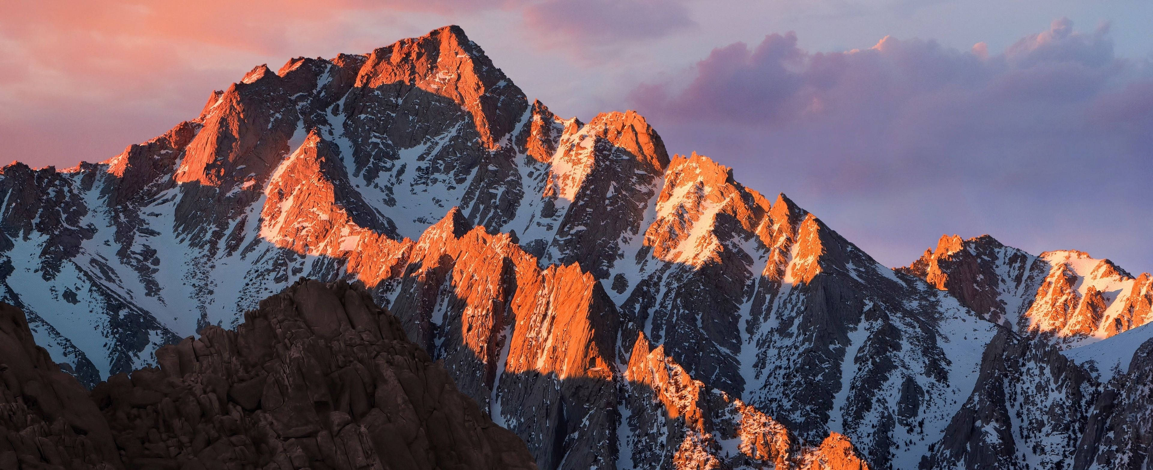 3840x1600 桌布 壁紙 — 夕陽將鋸齒狀的雪山山峰沐浴在溫暖的橙色光芒中，映襯著柔和的紫色天空。