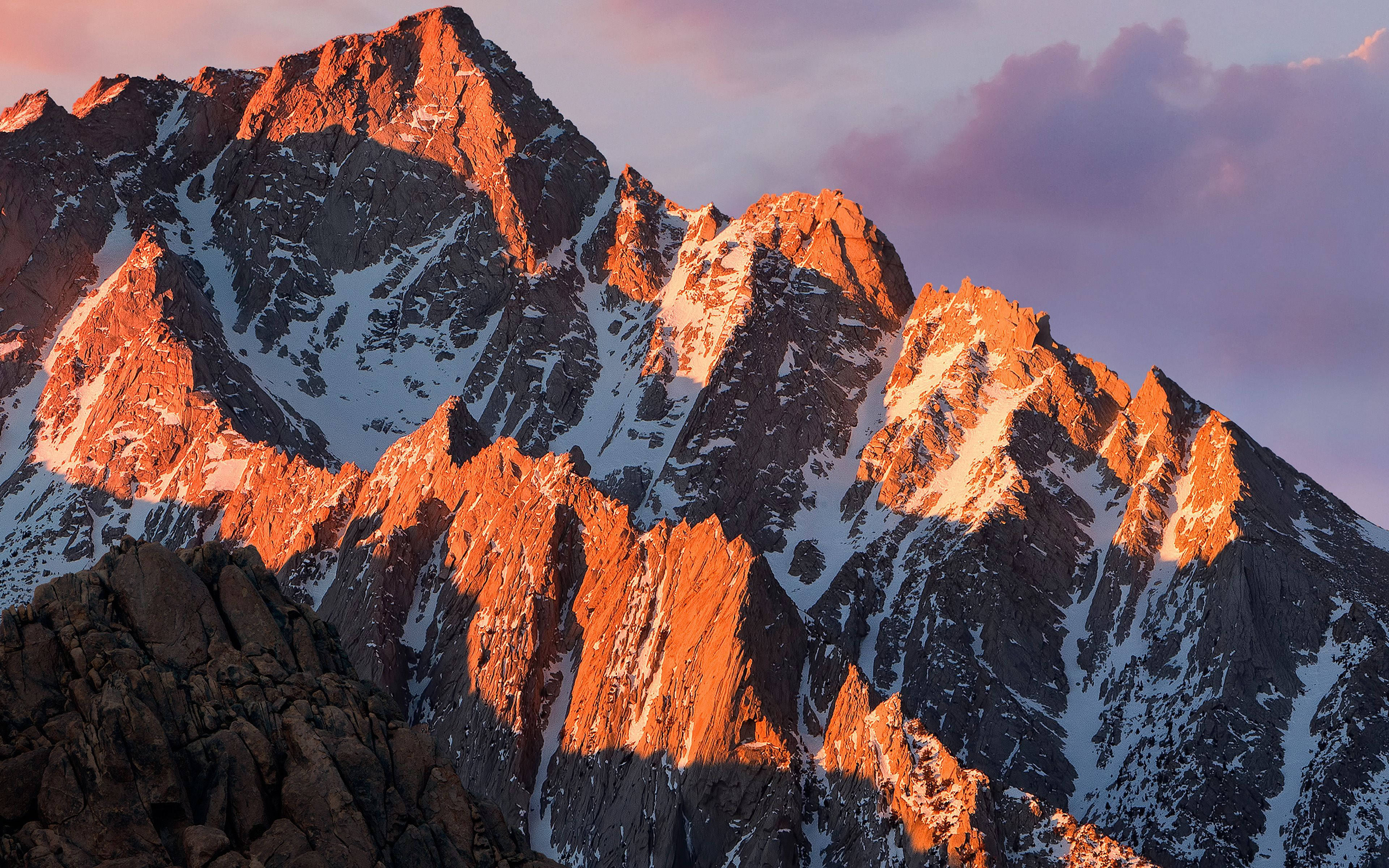 macOS 桌布 壁紙 — 夕陽將鋸齒狀的雪山山峰沐浴在溫暖的橙色光芒中，映襯著柔和的紫色天空。